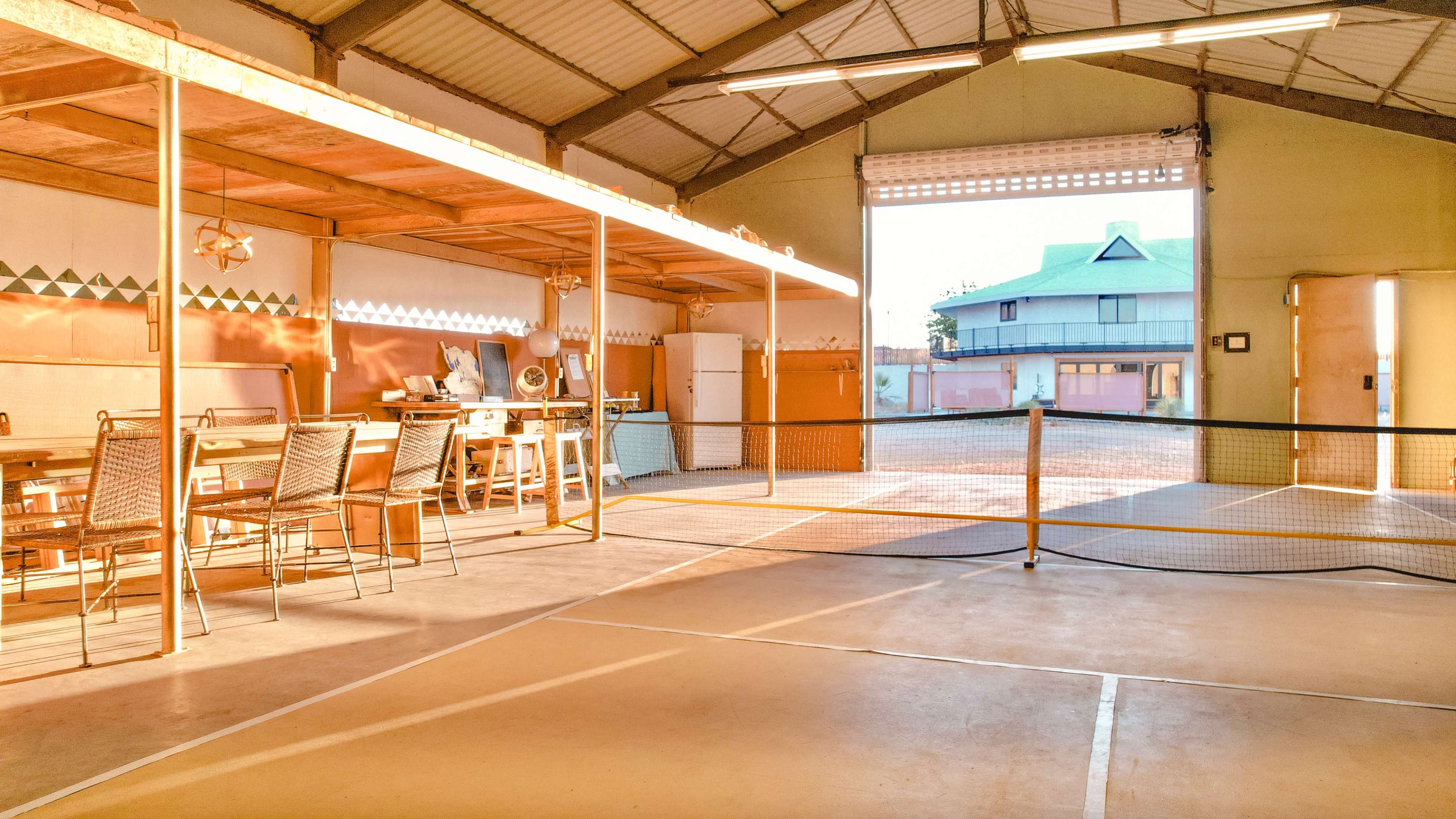The image shows an indoor space with a dining area and a view of an outdoor court through large open doors.