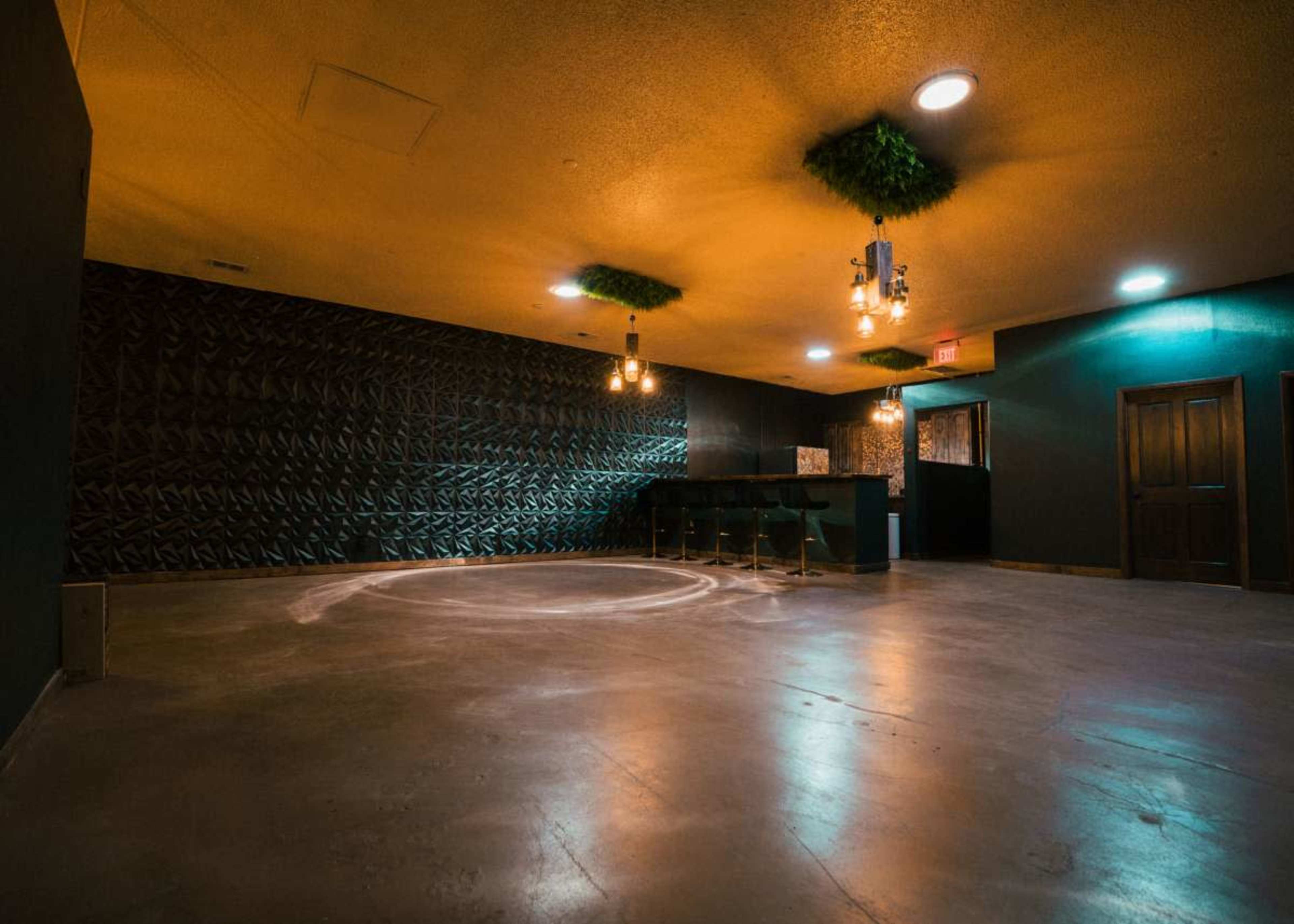 The image shows a spacious, dimly lit room with a dark textured wall, a bar area with stools, and several hanging light fixtures.