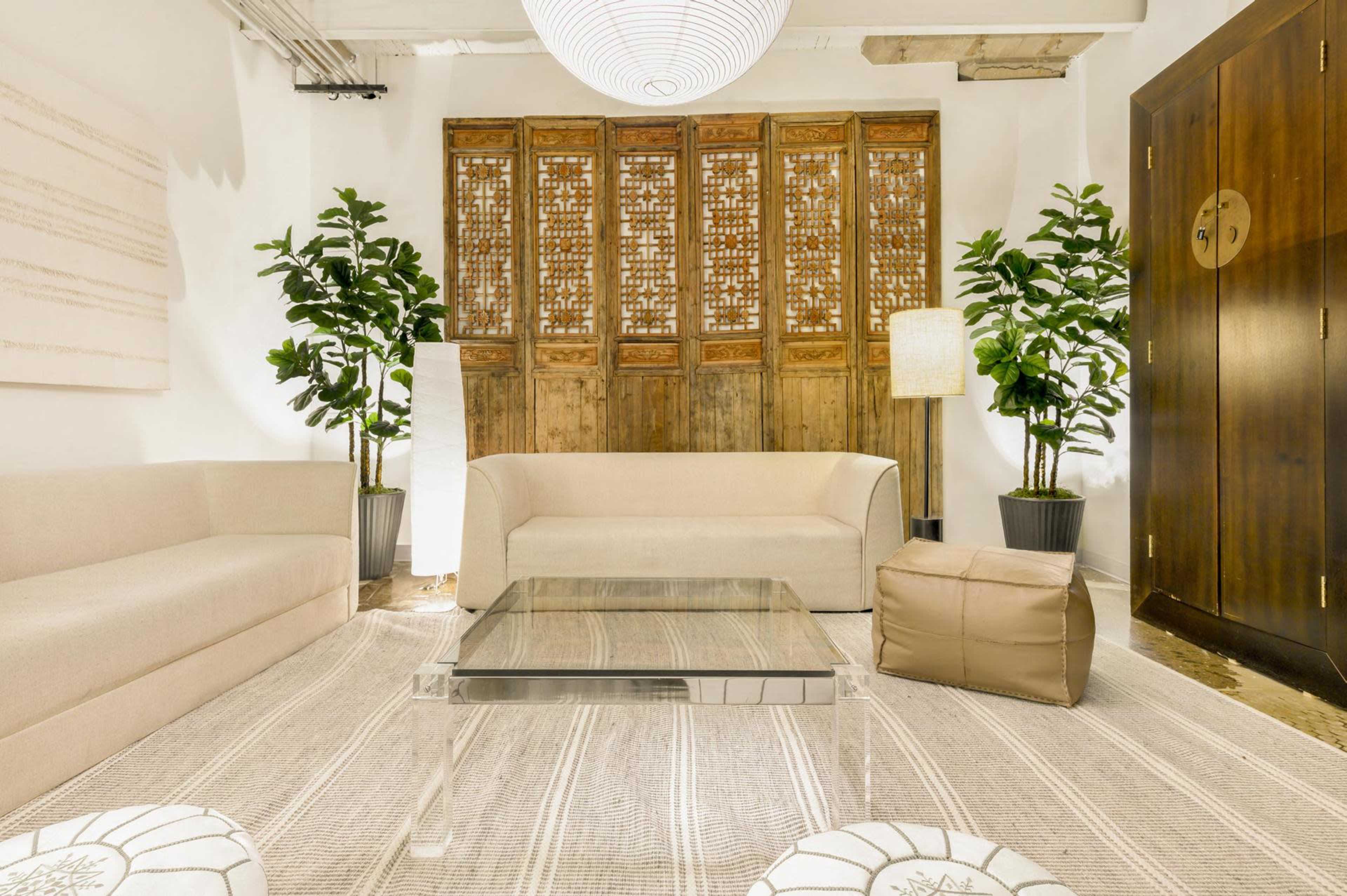 A modern living room features beige sofas, a glass coffee table, potted plants, and a wooden decorative screen against the wall.