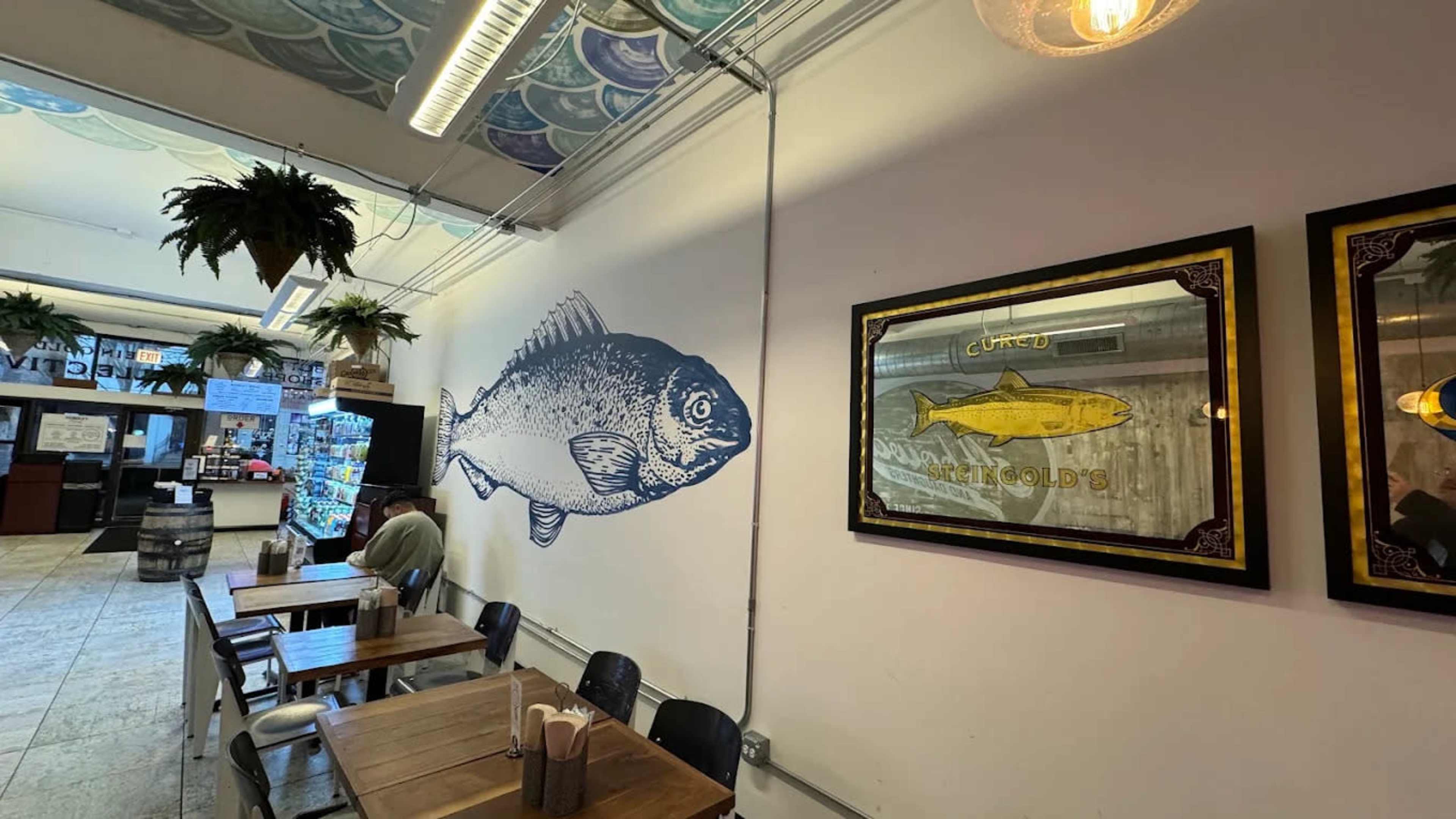 The image shows a restaurant interior featuring a large fish mural on one wall alongside framed art, with wooden tables and chairs arranged for dining.