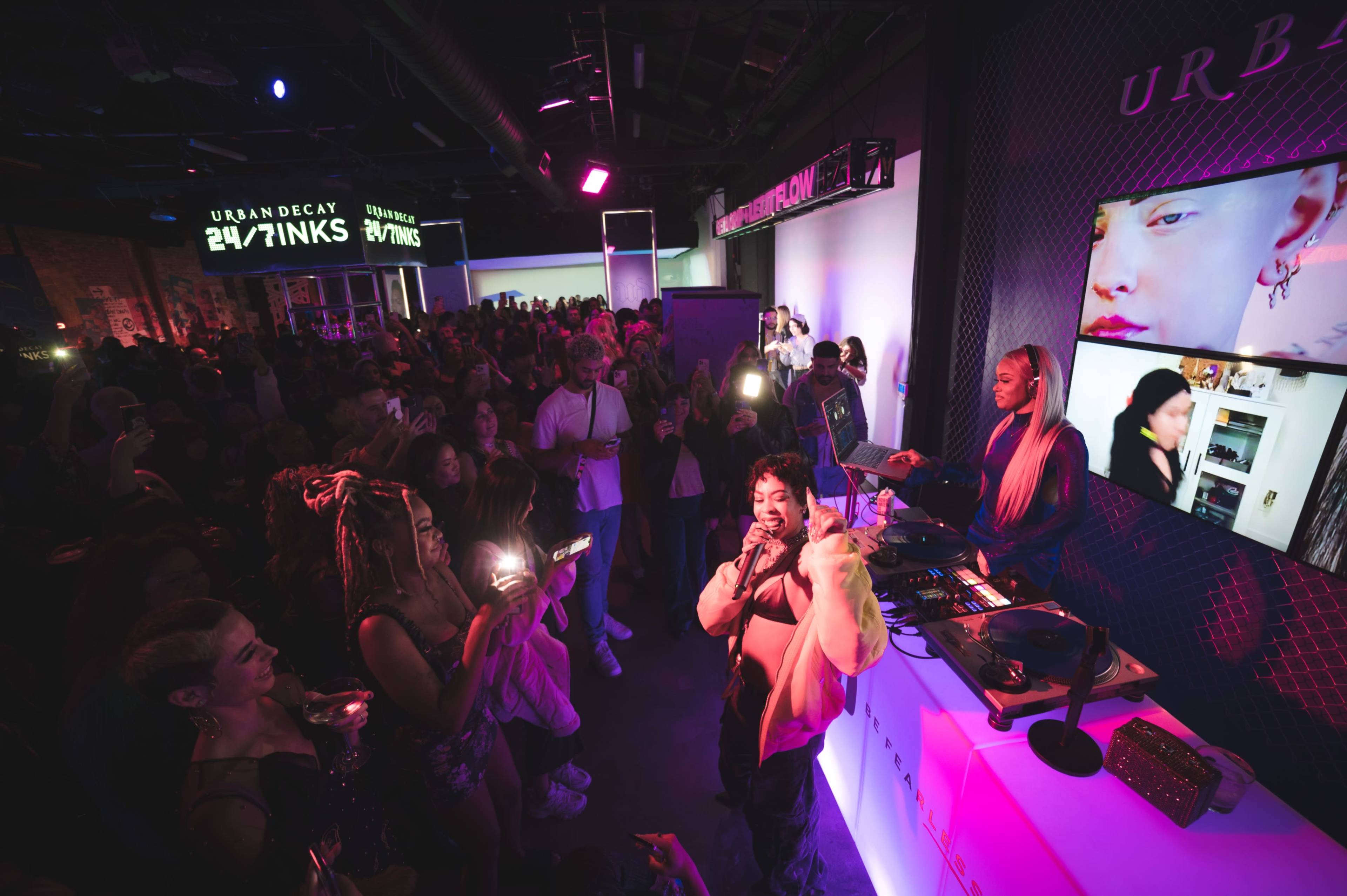 A crowded event features a performer on stage, surrounded by an audience, with colorful lights and large screens displaying visuals in a lively atmosphere.