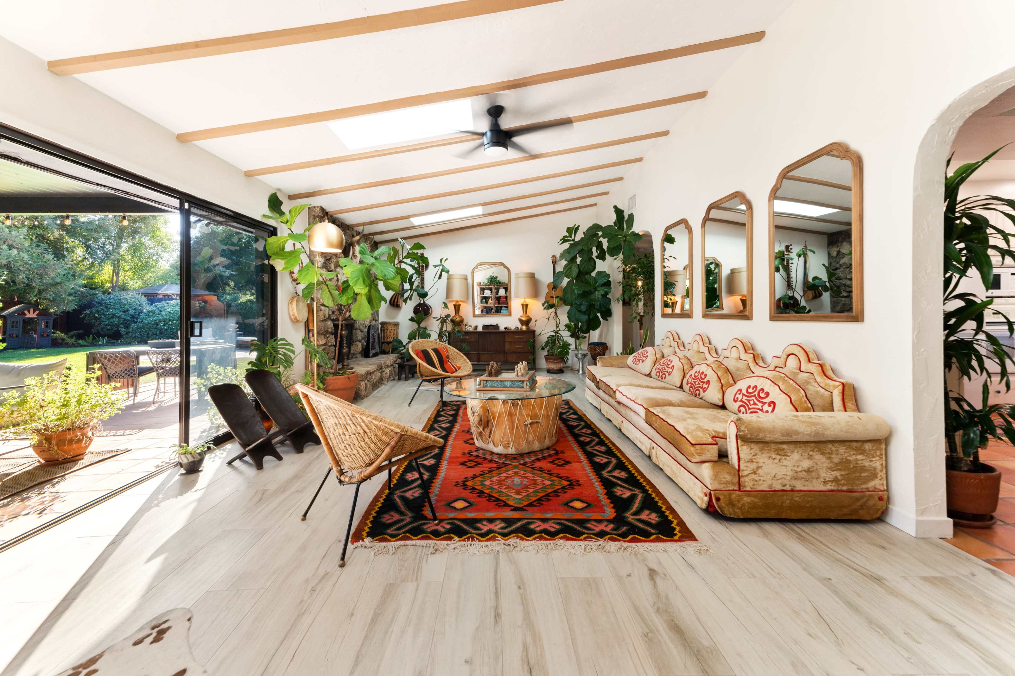 A bright living room with large windows, featuring a mix of plants, a patterned area rug, and comfortable seating arrangements.