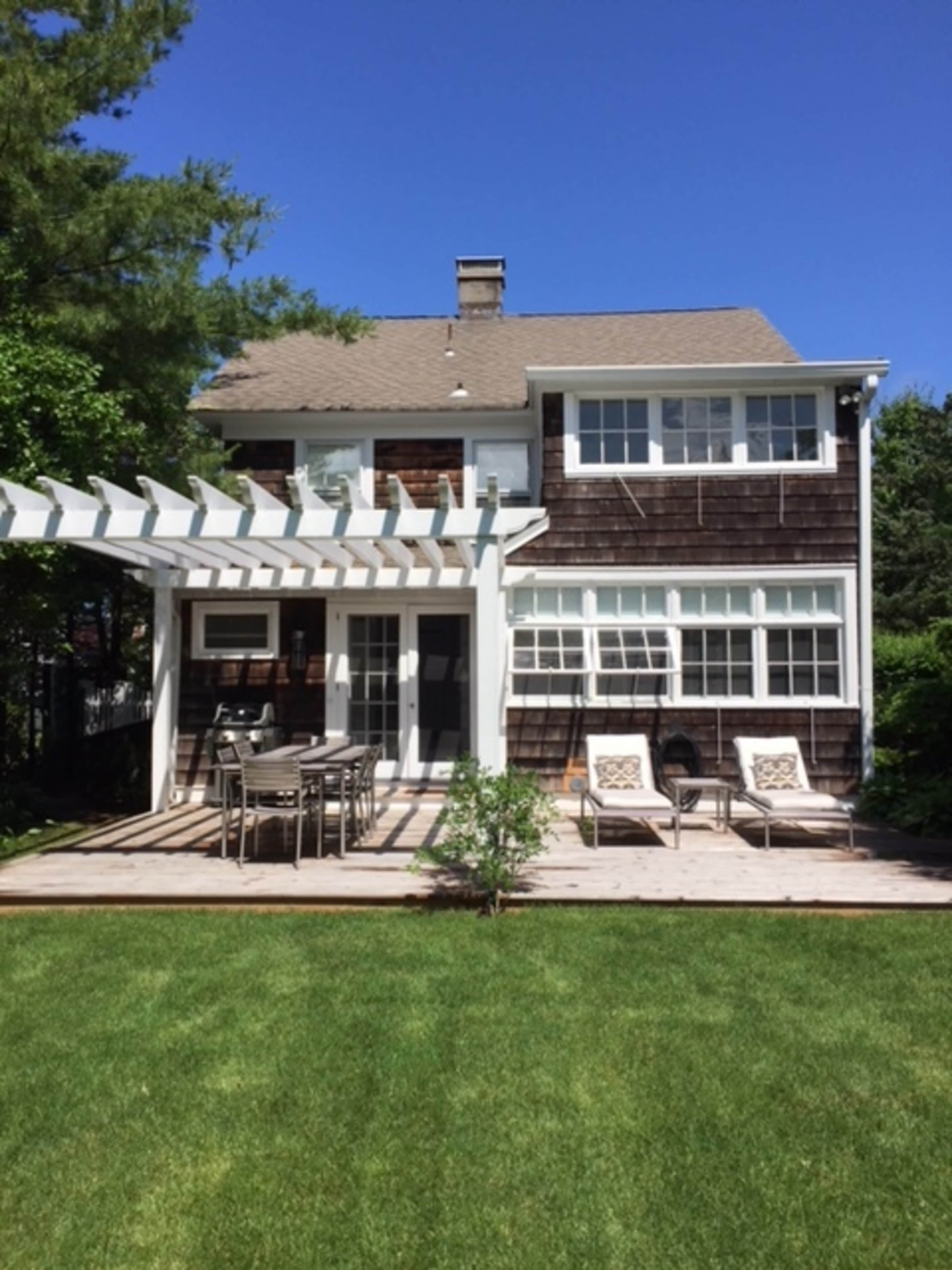 A two-story house features a wooden deck with outdoor furniture and a pergola, surrounded by a grassy lawn and trees.