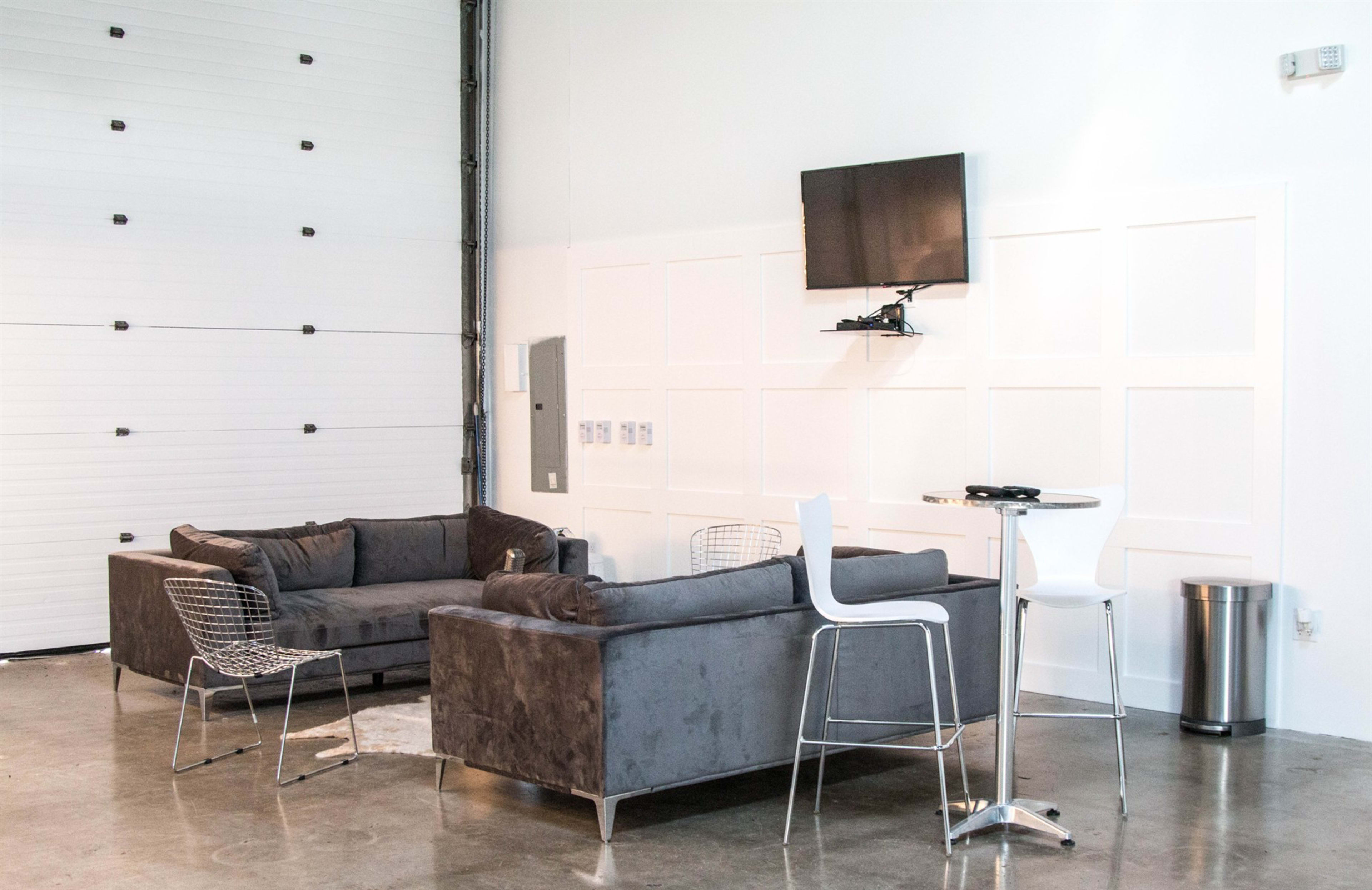 The image shows a modern lounge area with gray sofas, white chairs, a small table, and a wall-mounted television in a spacious, bright room.