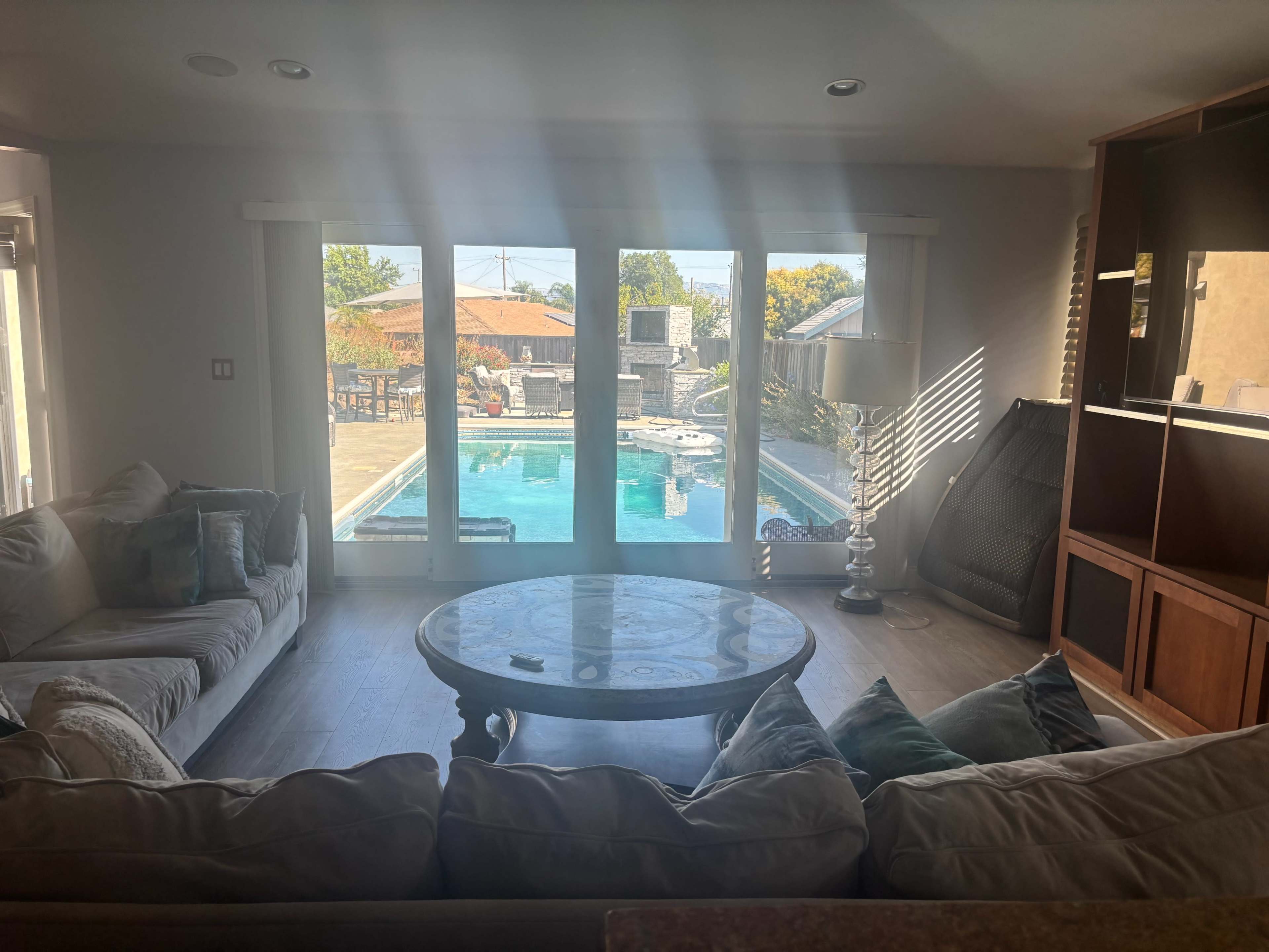 The image shows a cozy living room with a large sectional sofa, a round coffee table, and large windows that open to a pool area outside.
