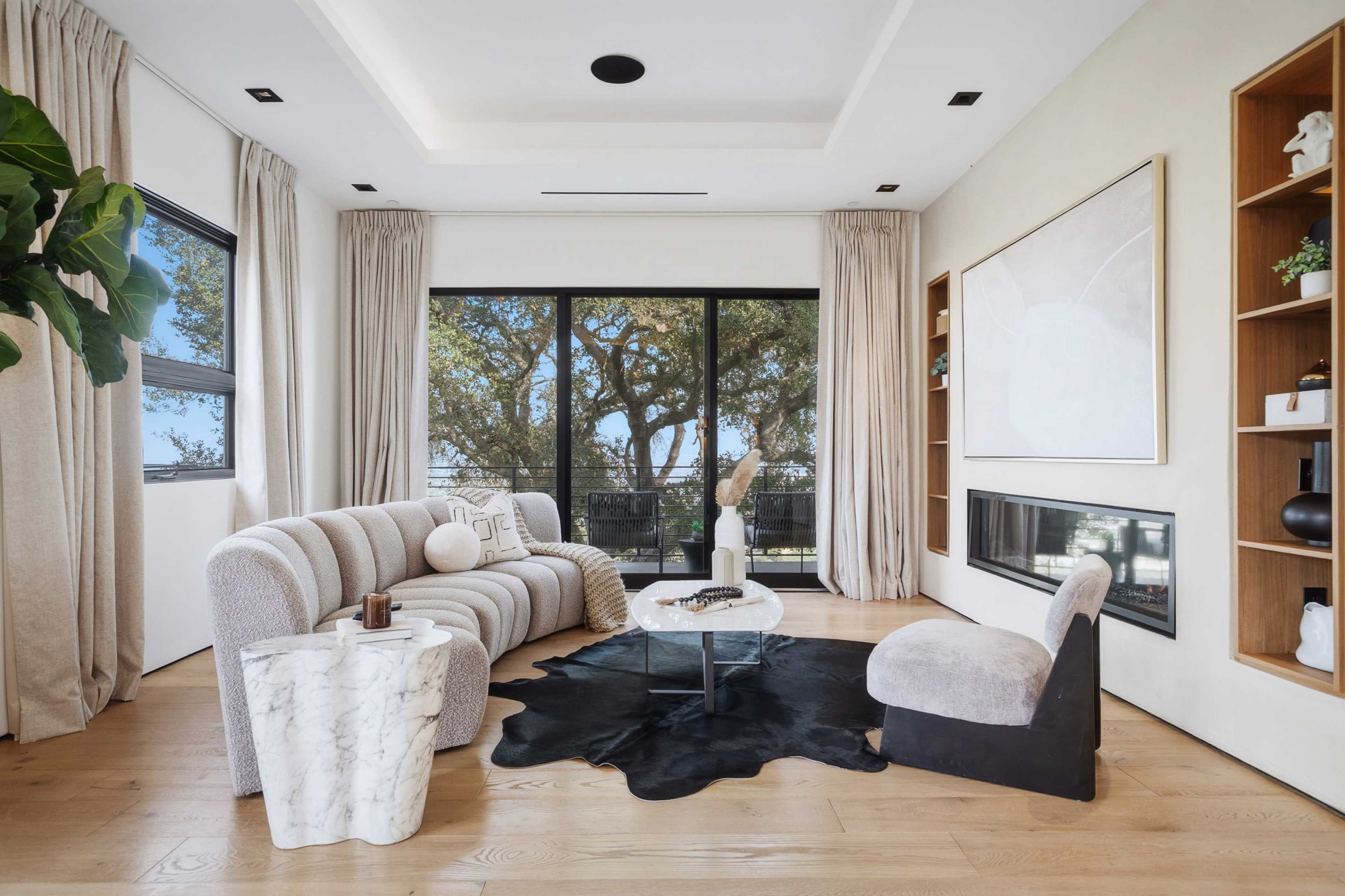 A modern living room features a curved sofa, a round coffee table, a fireplace, and large windows with views of trees outside.