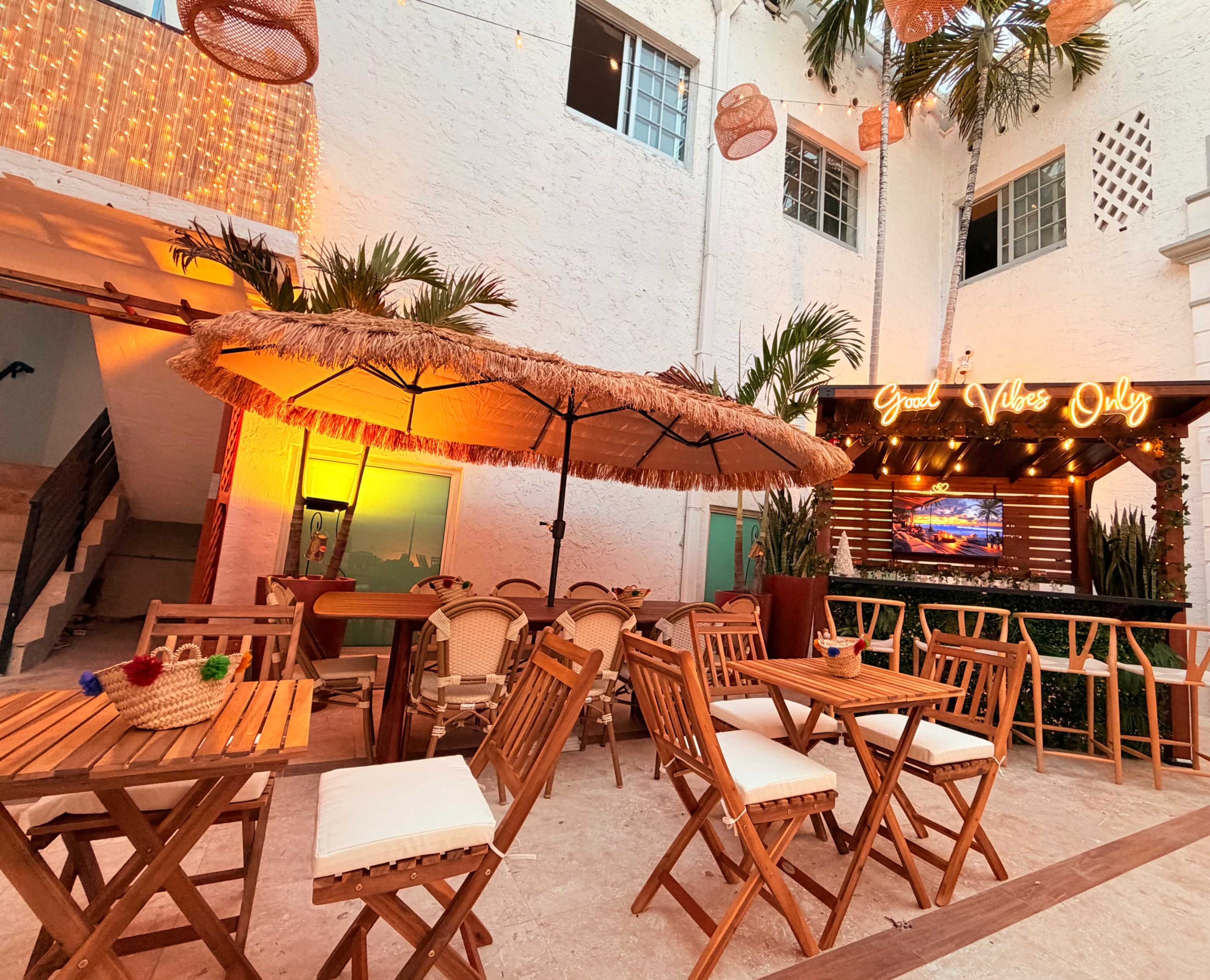 The image shows a cozy outdoor seating area with wooden tables and chairs, a large umbrella, and decorative lights reflecting a warm ambiance.