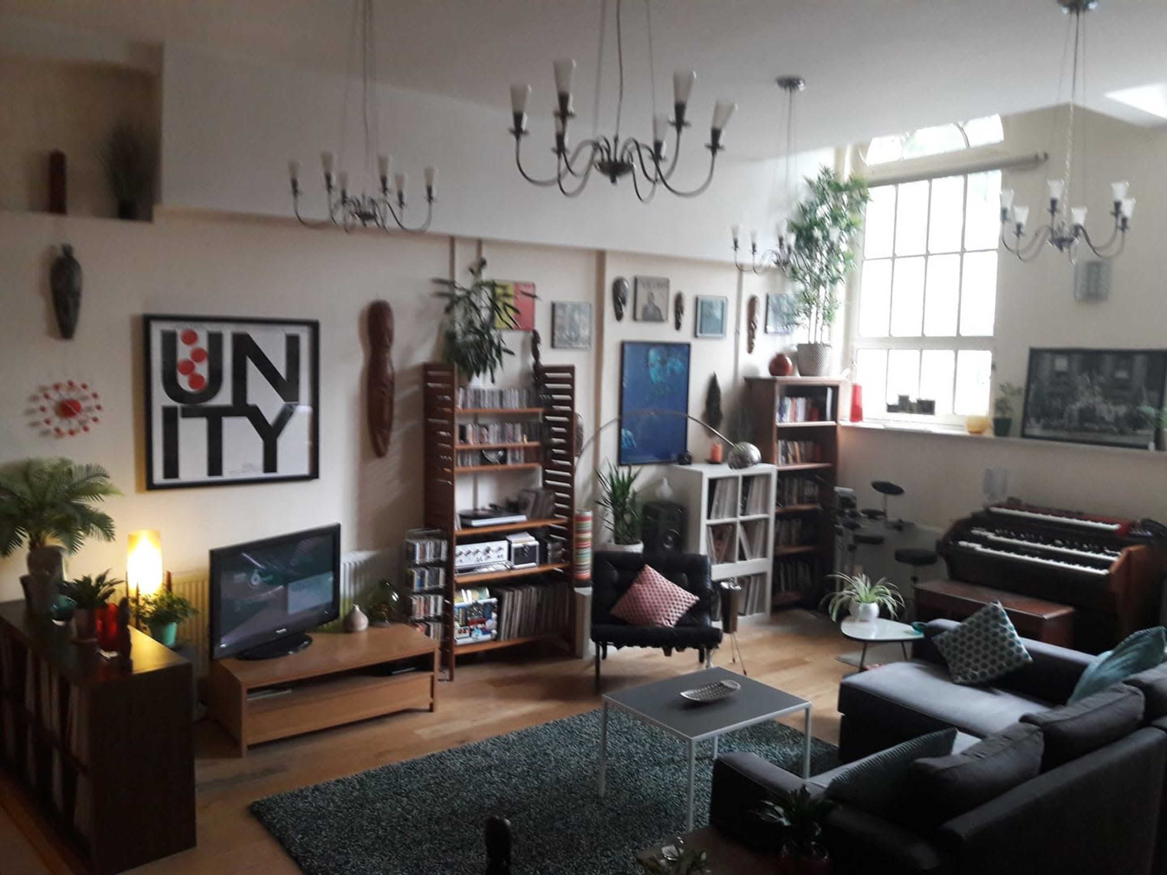 The image shows a spacious living room with high ceilings, featuring a mix of furniture, shelves filled with books, plants, and a keyboard placed against the wall.