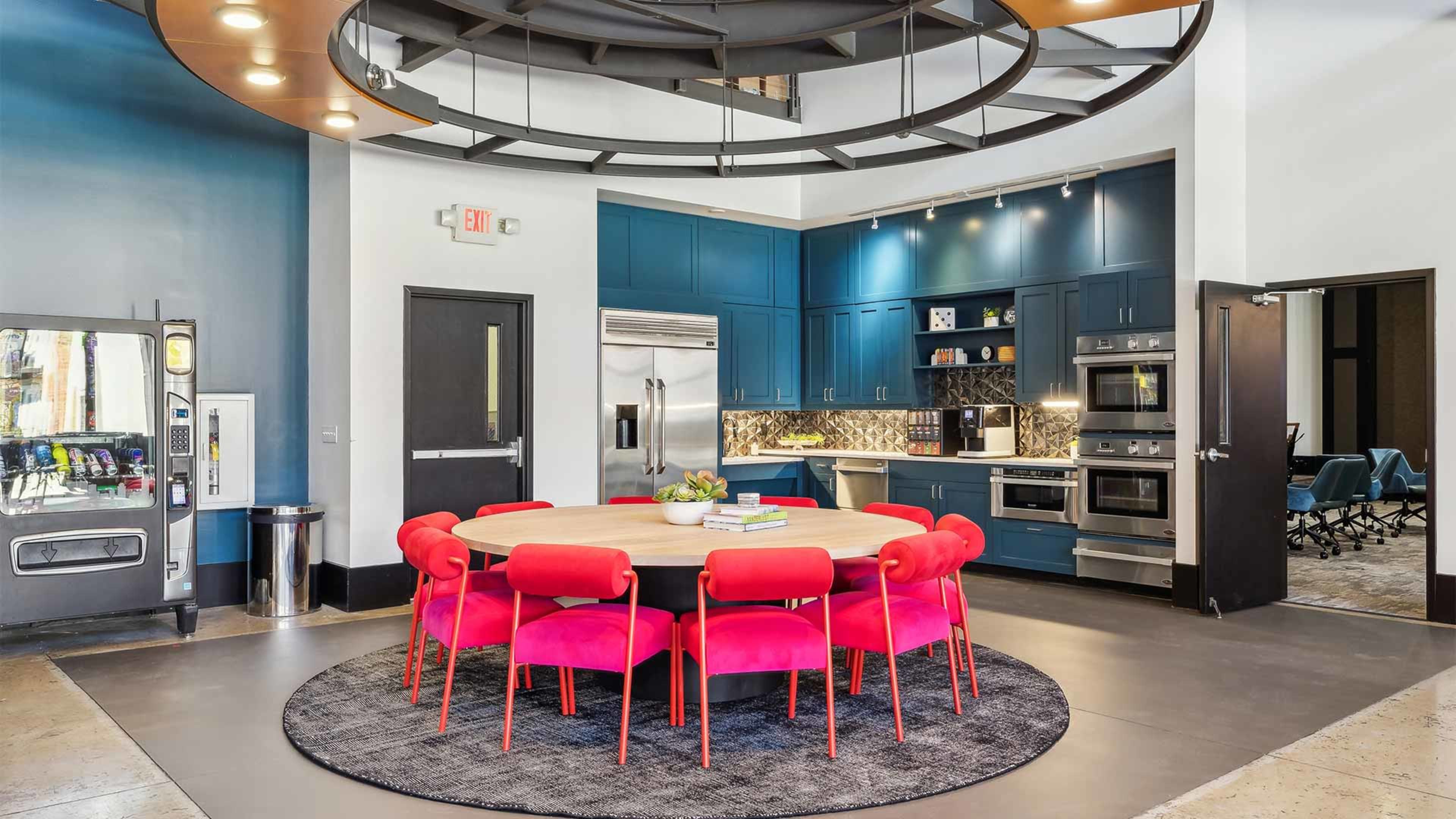 A modern kitchen features a round wooden table surrounded by bright pink chairs, with dark blue cabinetry and appliances in the background.