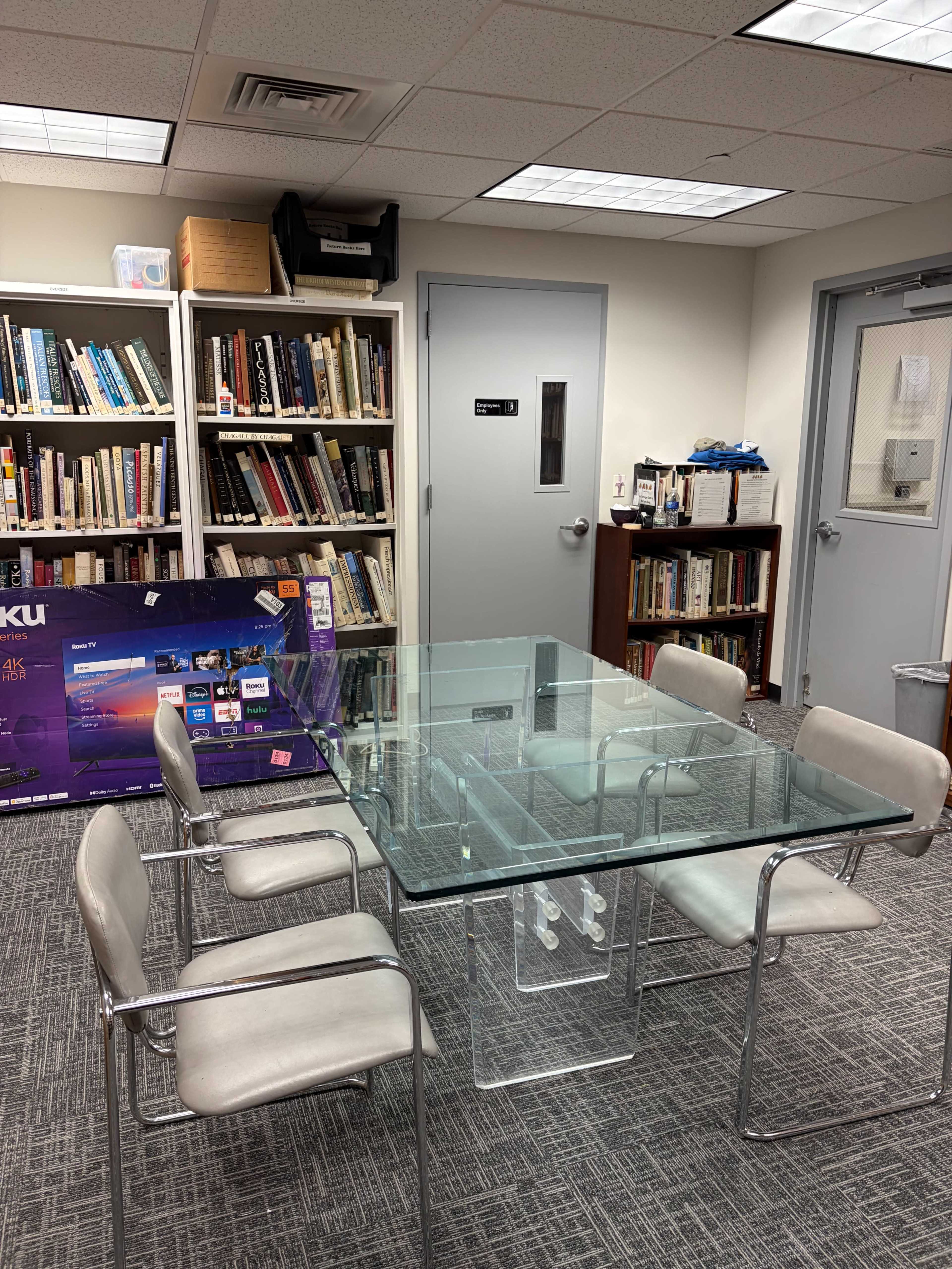 The image shows a glass conference table surrounded by four chairs, with a bookshelf in the background and a Roku device on display.