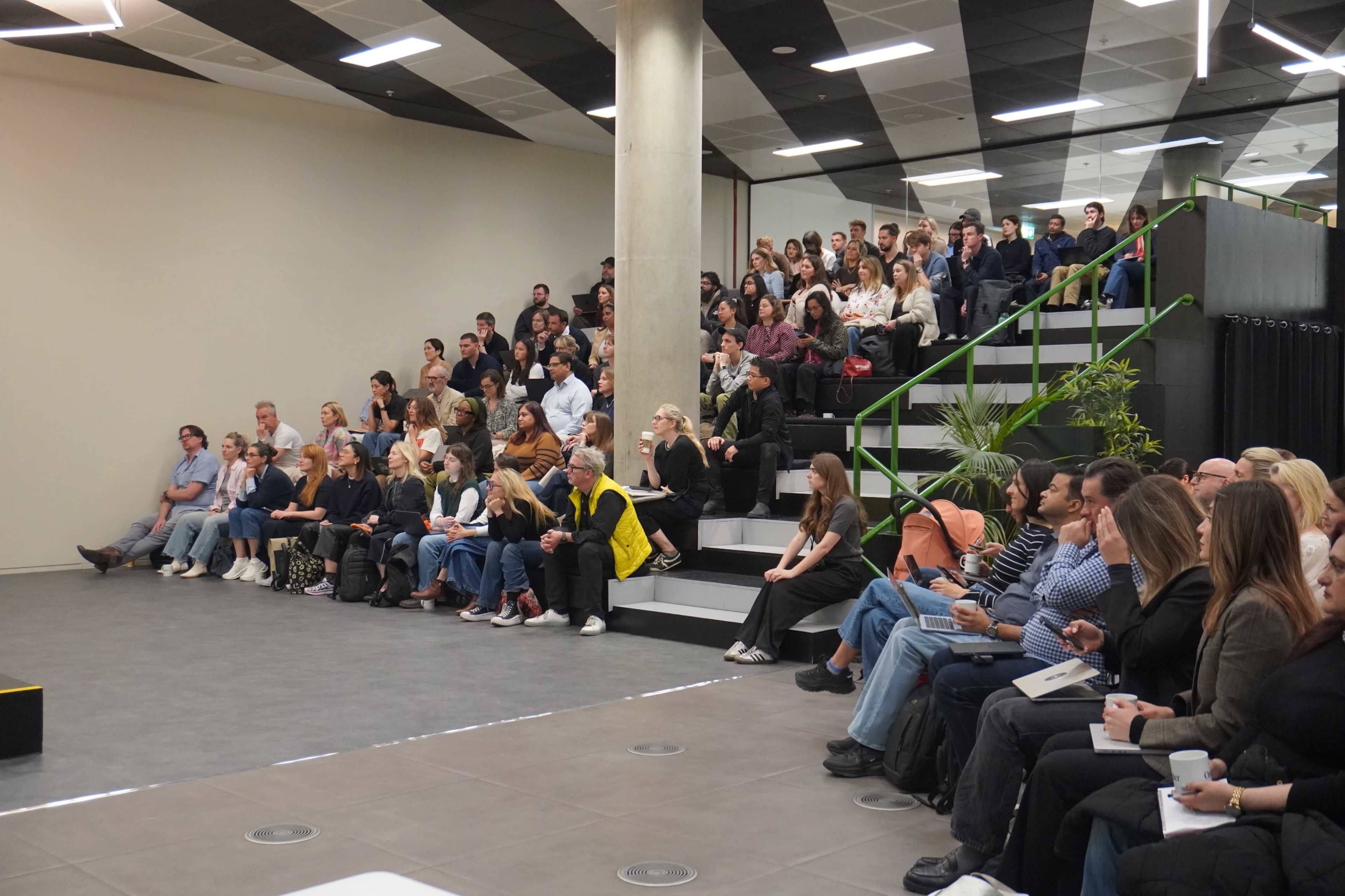 A large audience is seated in a modern auditorium, attentively listening to a speaker on a stage.