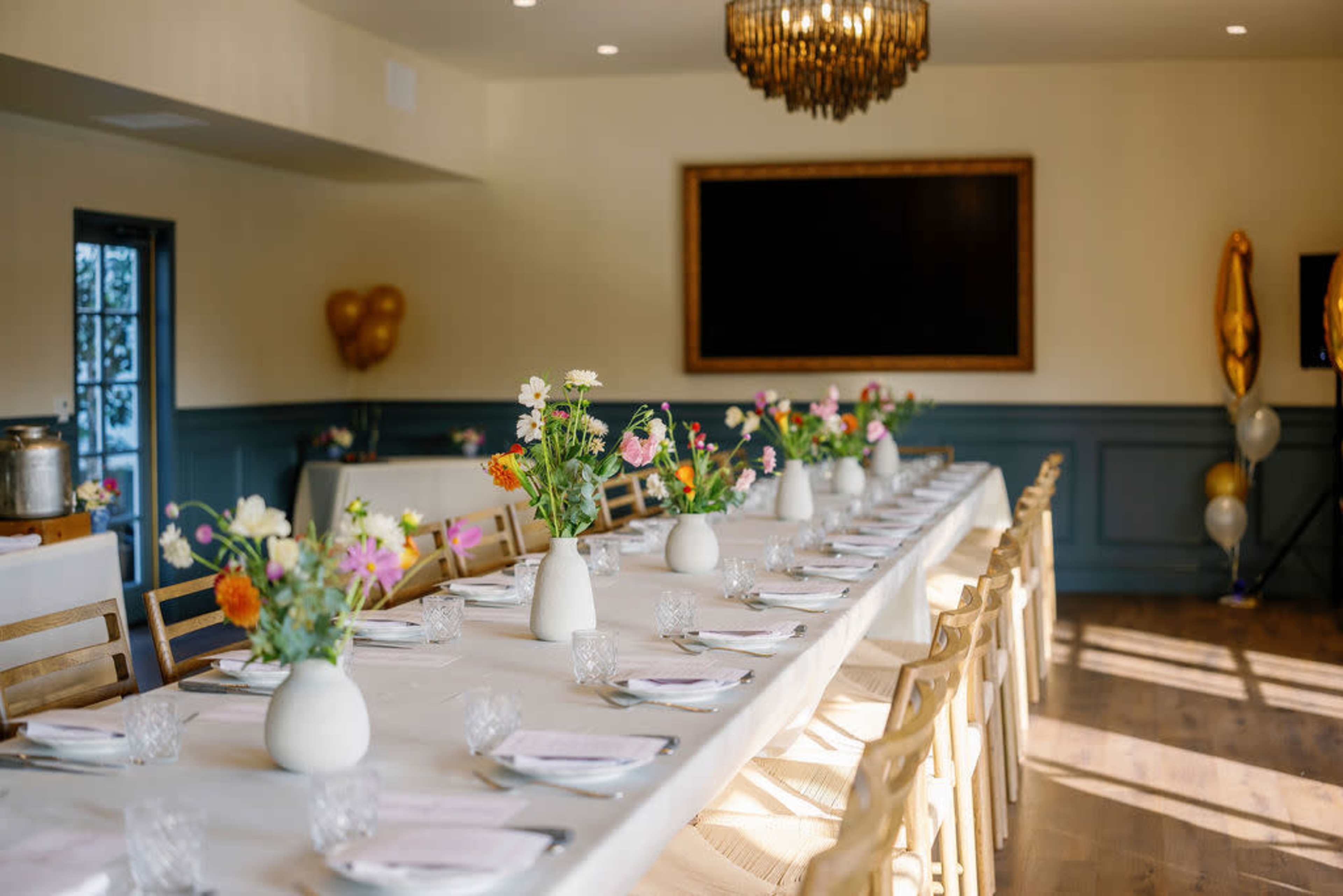A long dining table is set with white tablecloths and vases of flowers, illuminated by a chandelier in a well-lit room.
