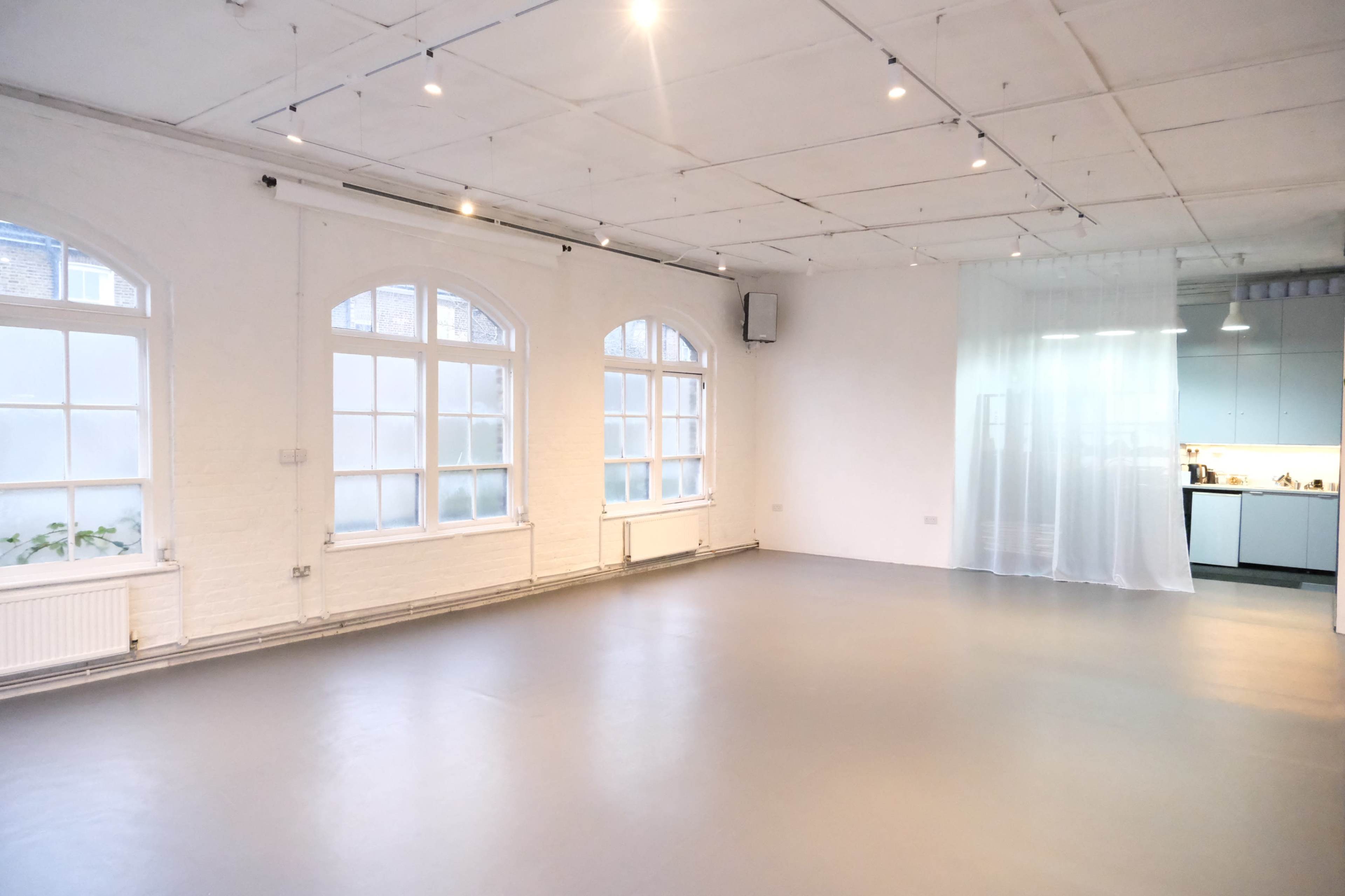 The image shows a spacious, well-lit interior room with white brick walls, large windows, and a partial view of a kitchen area behind a sheer curtain.