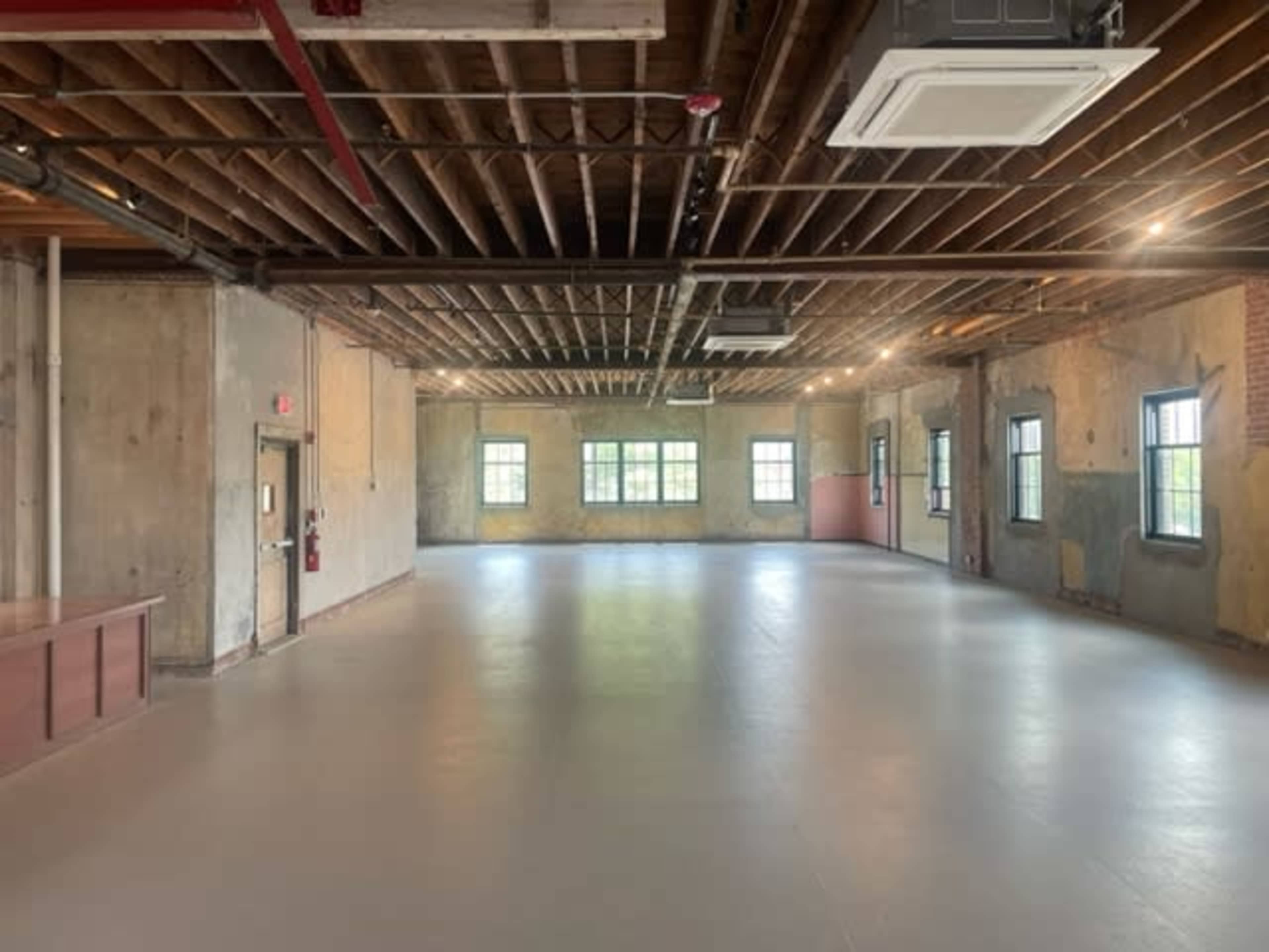 A spacious, empty room with exposed wooden beams, concrete walls, and several windows.