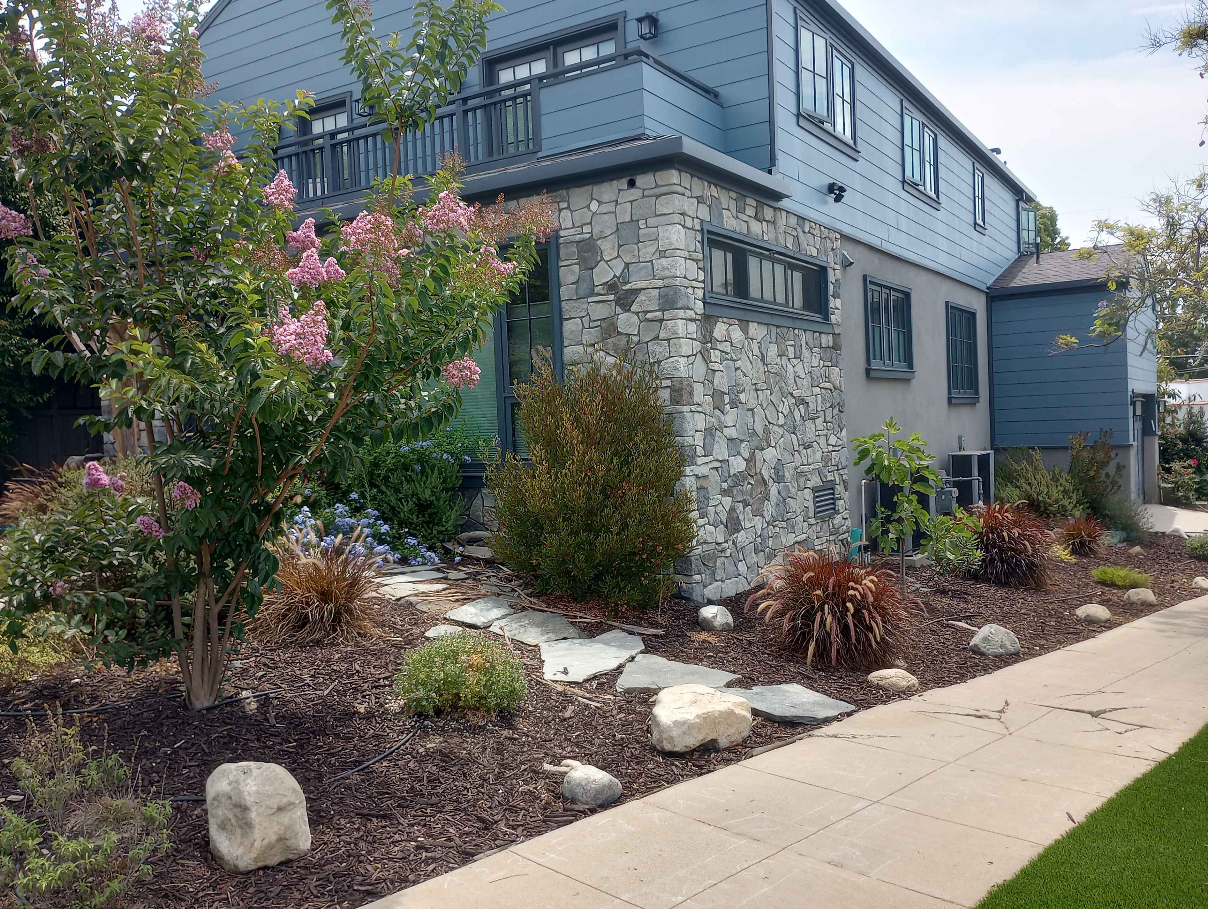 A modern house with a stone and wood exterior, surrounded by landscaped gardens featuring various plants and stone pathways.