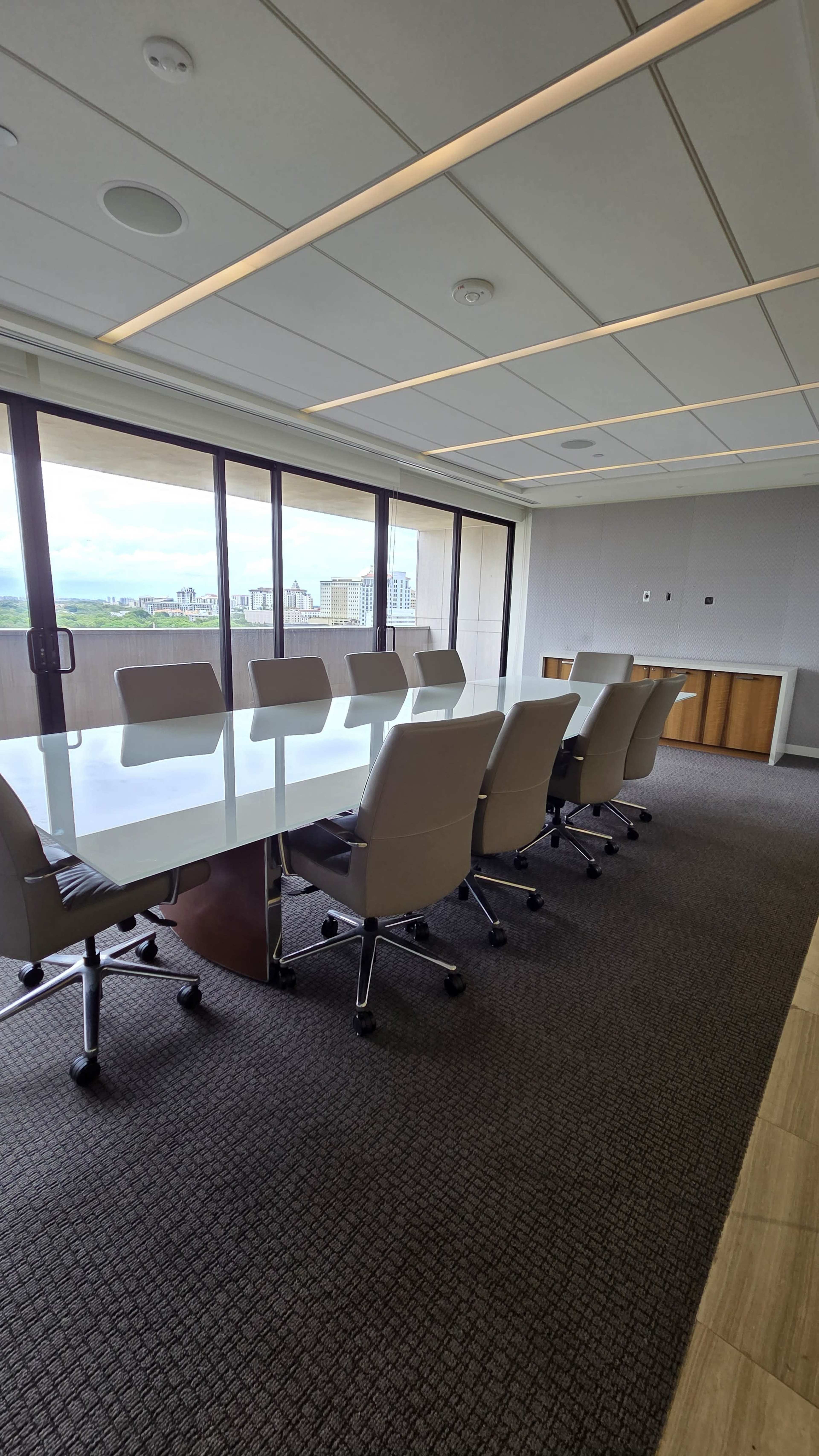 The image shows a modern conference room with a long white table, ergonomic chairs, and large windows overlooking a cityscape.