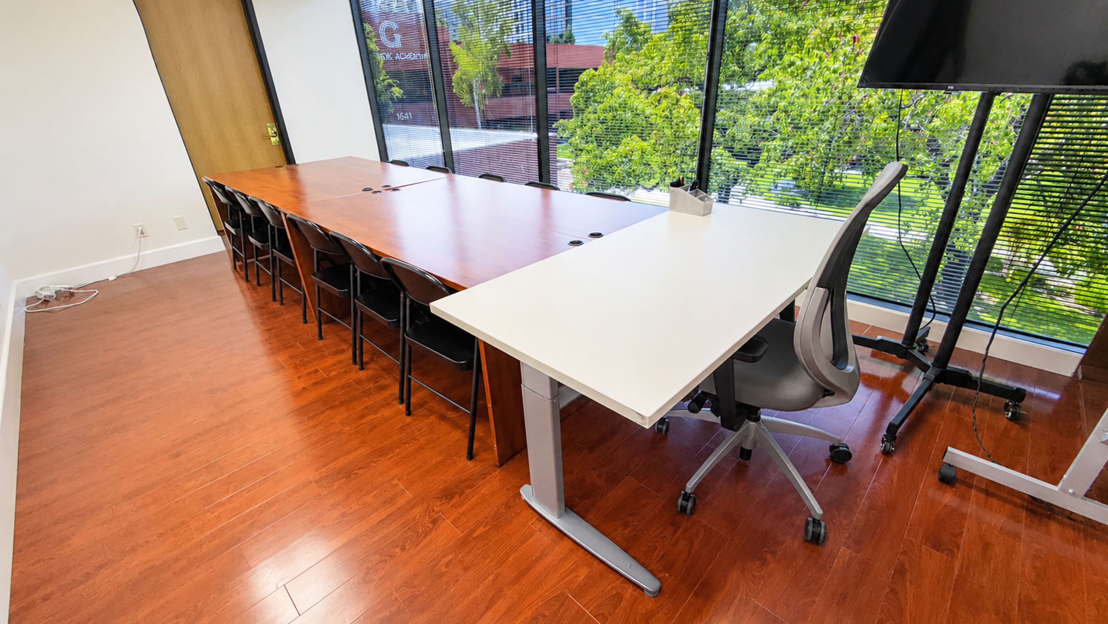 The image shows a modern conference room with a long wooden table and several black chairs facing large windows, along with a white desk and a rolling chair in the corner.