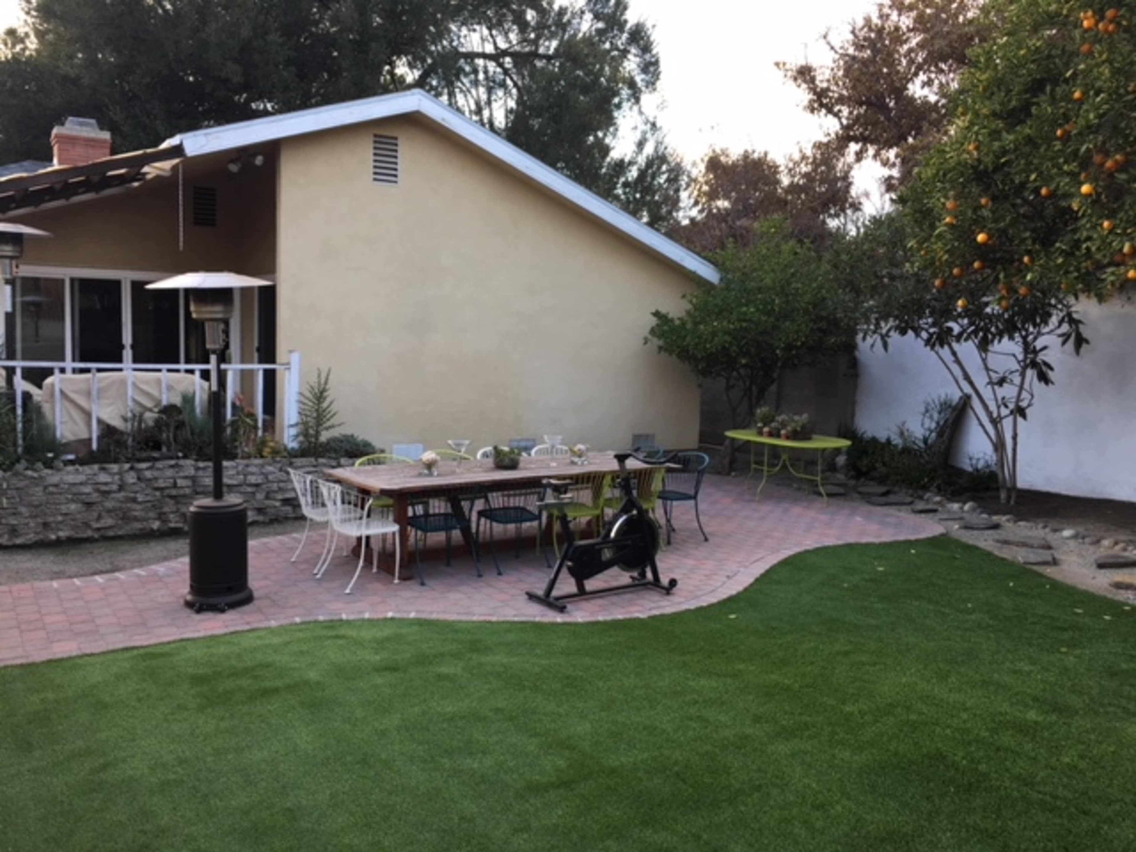 A backyard scene features a paved patio with various outdoor furniture, including tables and chairs, surrounded by greenery and an orange tree.