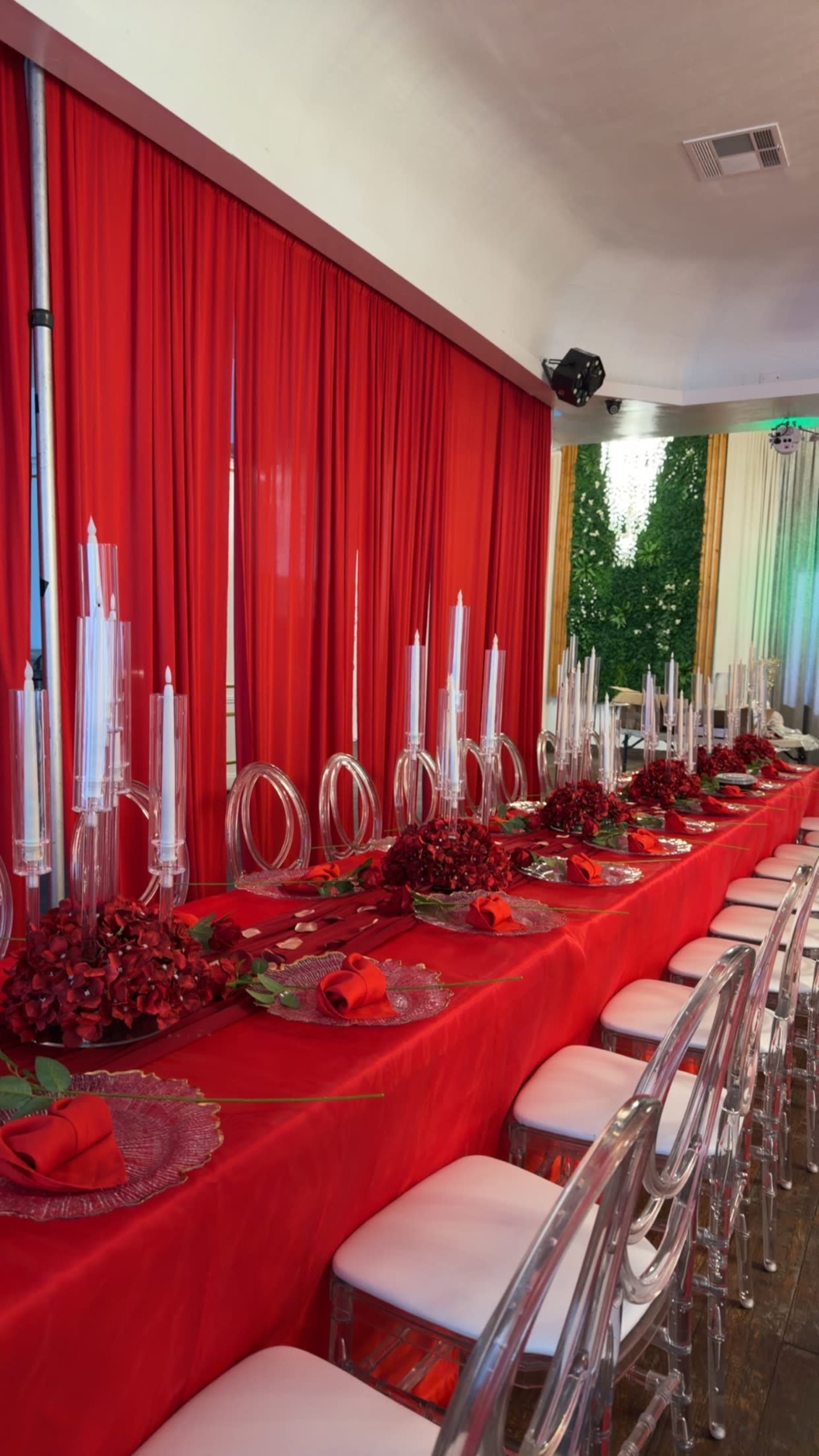 A long table is set with red tablecloths, crystal candlesticks, and decorative arrangements of roses surrounded by clear acrylic chairs.