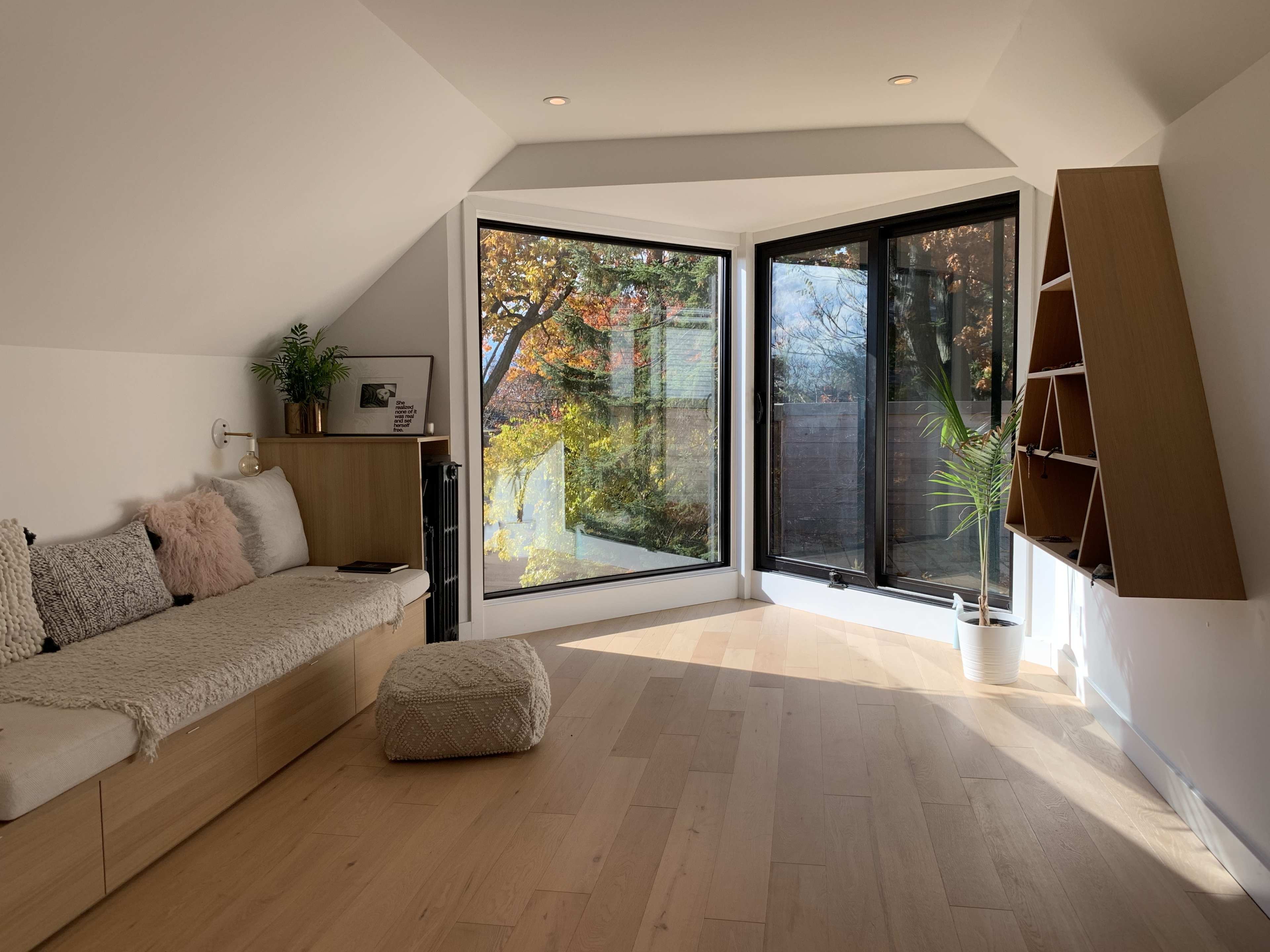 The room features a cozy seating area with a cushion, large windows showcasing a view of trees, and a decorative plant in the corner.