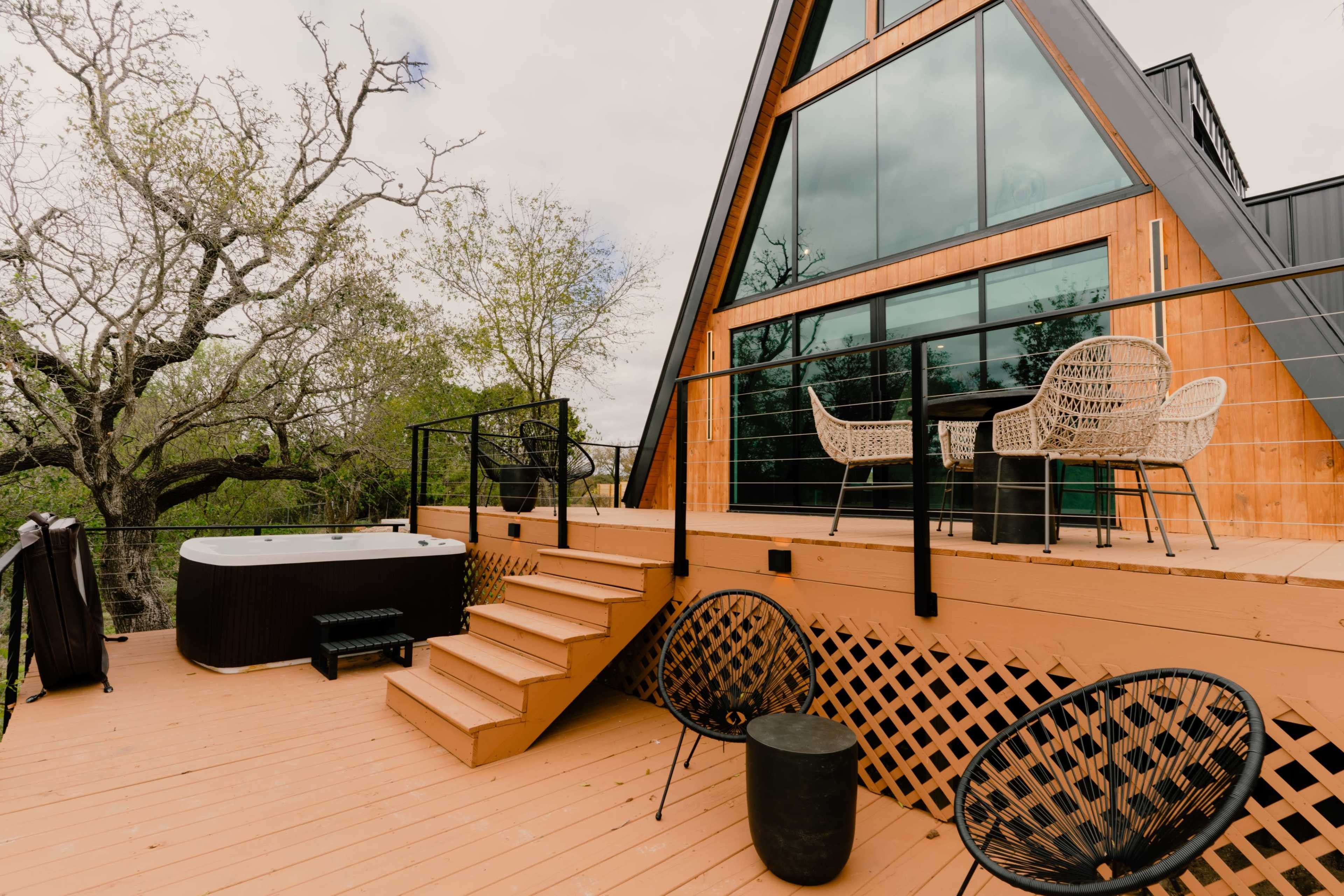 A modern A-frame house with large windows, a hot tub, and a wooden deck surrounded by trees.
