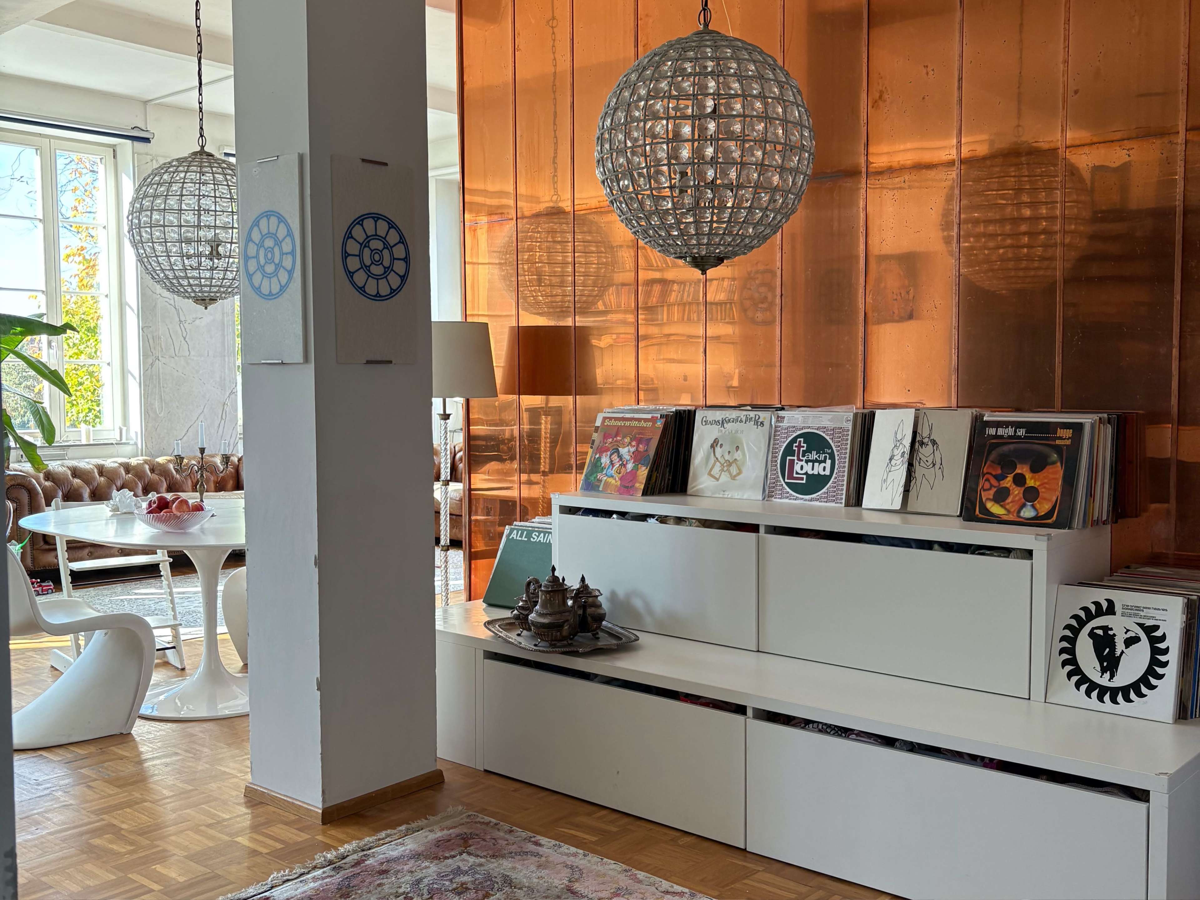 The image shows a modern interior with a white shelving unit displaying vinyl records, a round dining table, and large pendant lights against a copper-colored wall.