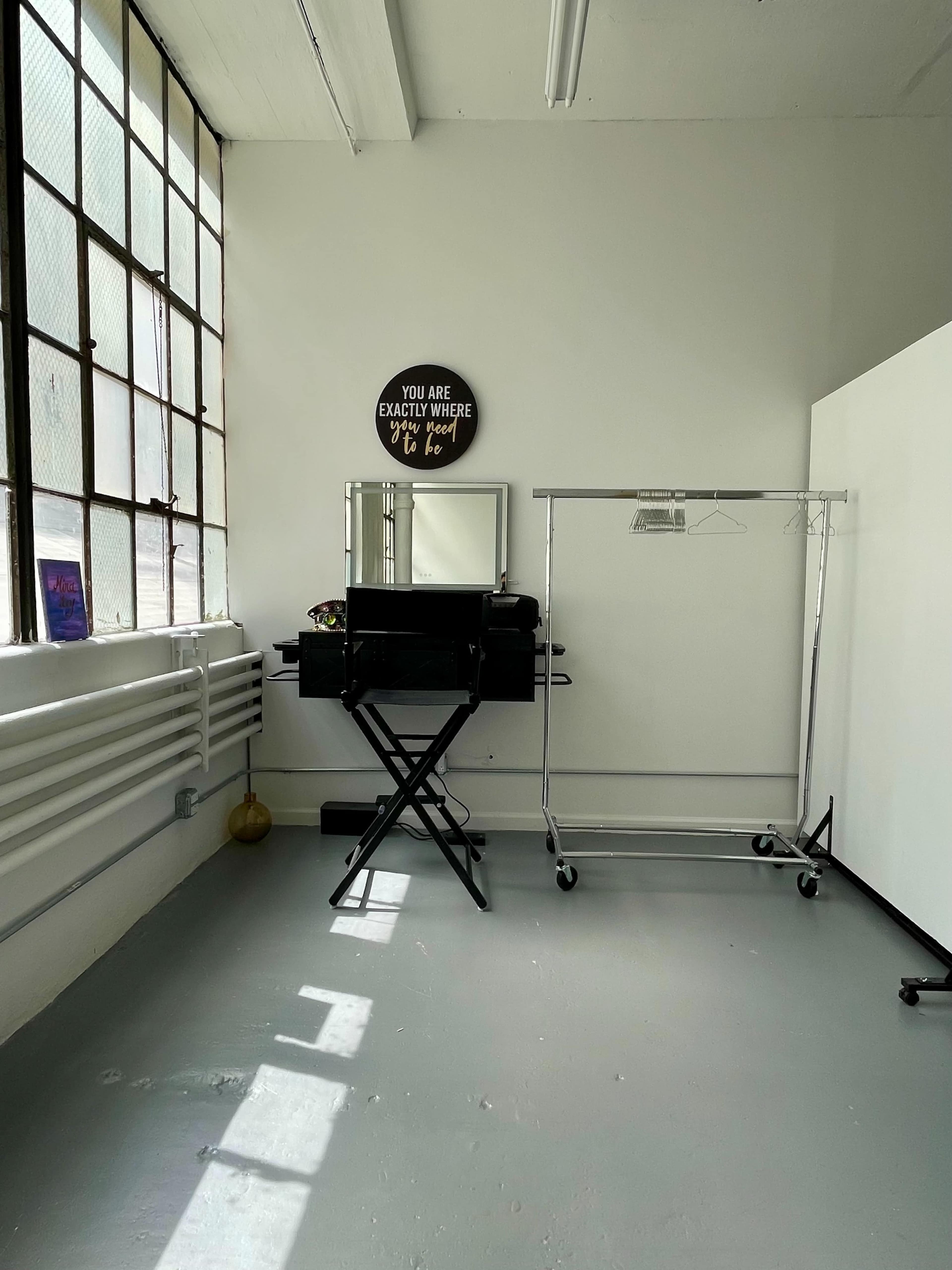 The image shows a minimalistic interior space with large windows, a mirror on a black stand, a clothing rack, and a sign on the wall.
