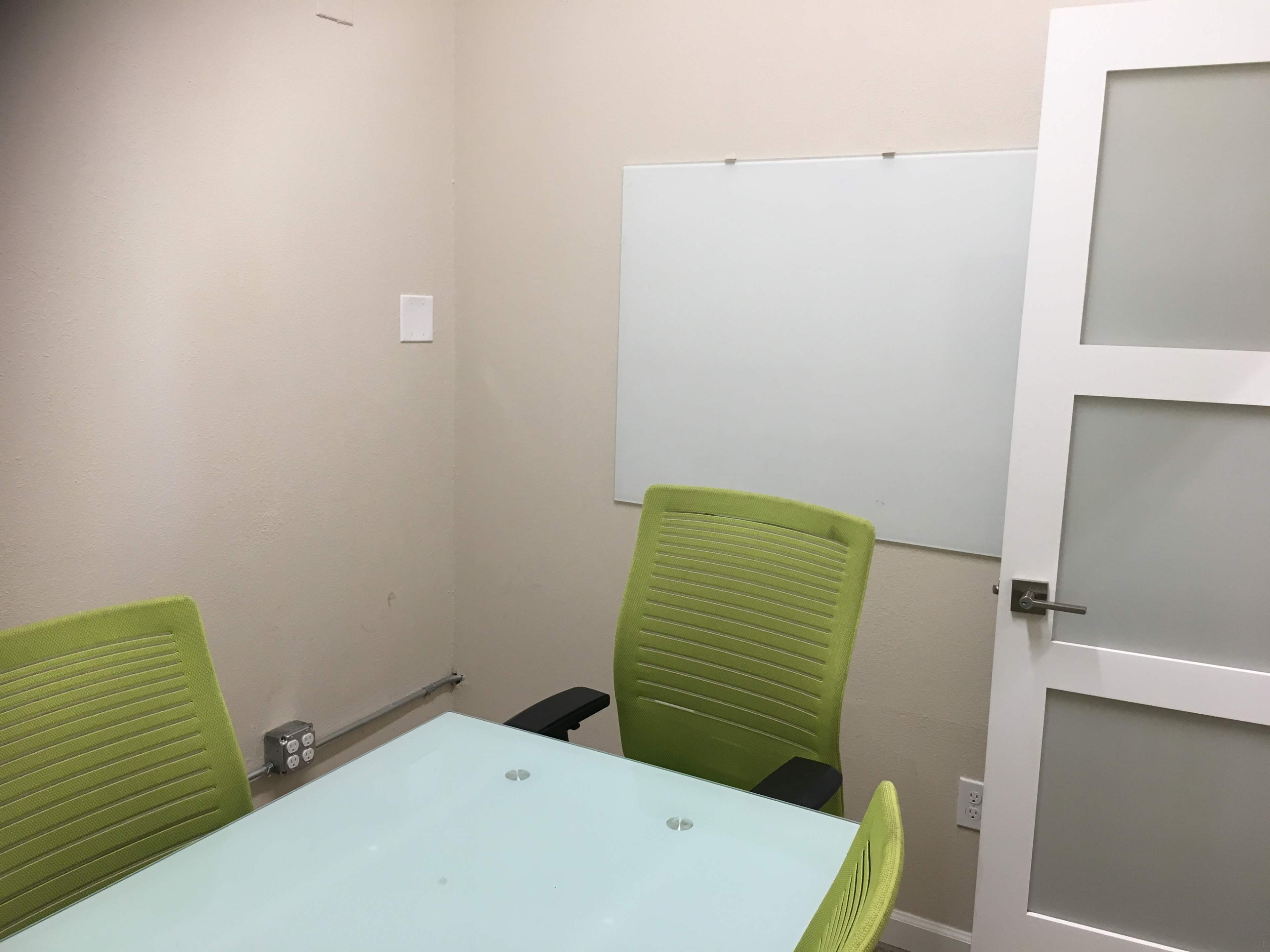 The image shows a small conference room with a glass table, two green chairs, and a whiteboard on the wall.