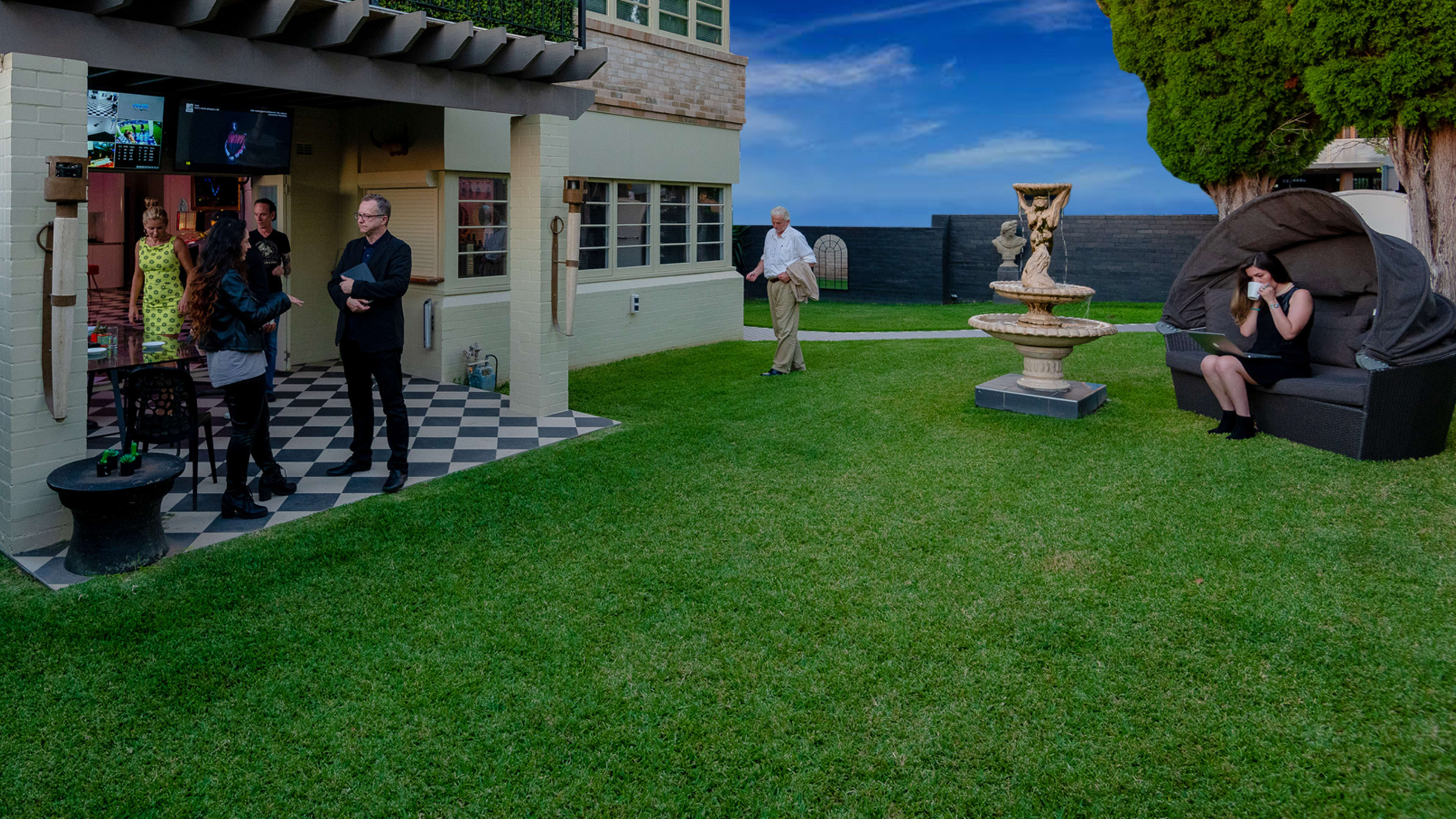 A gathering in a well-manicured lawn with a group of people socializing around a house, a fountain, and a large chair.