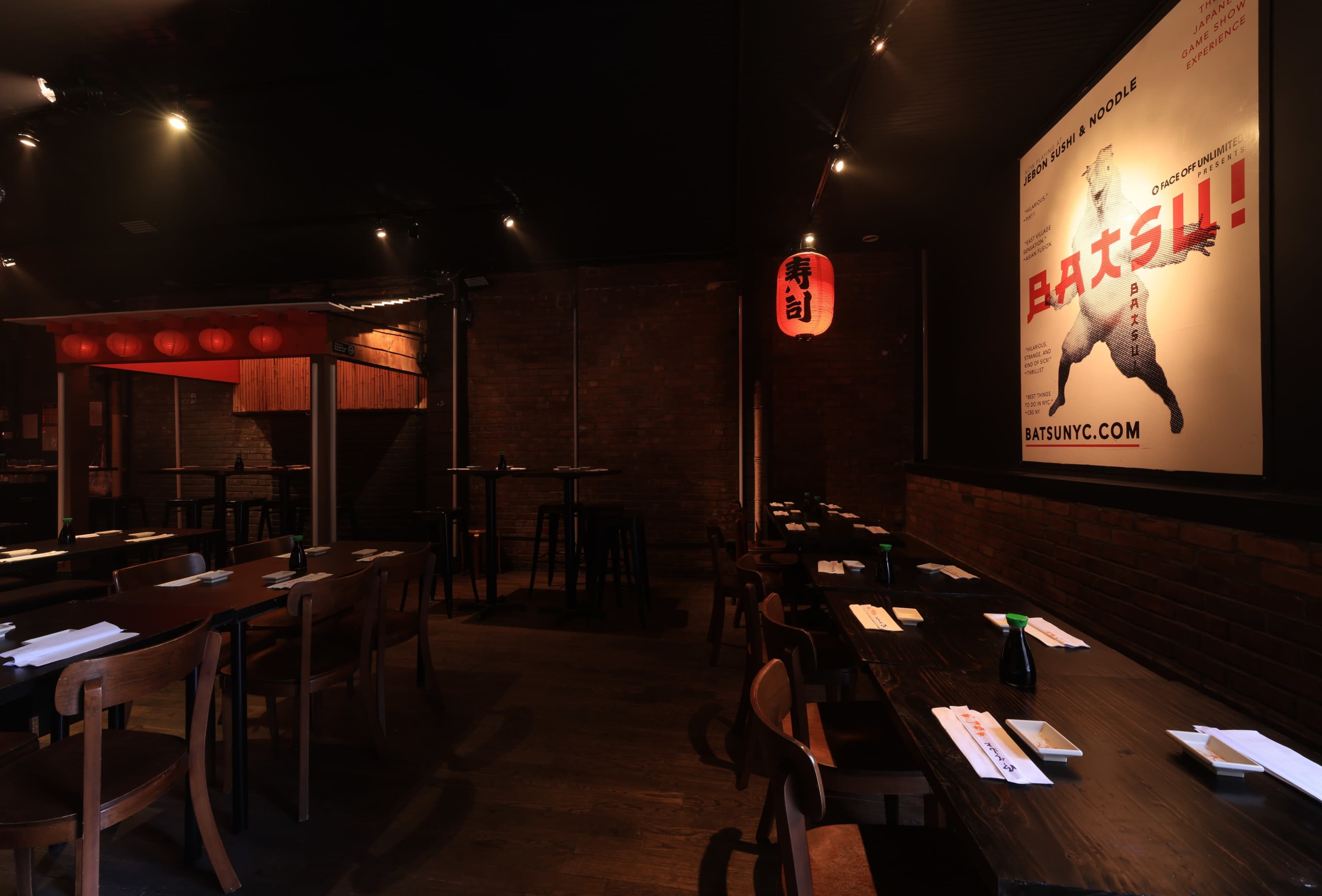 An empty restaurant interior with dark wooden tables, chairs, and red lanterns, featuring a large poster on the wall.