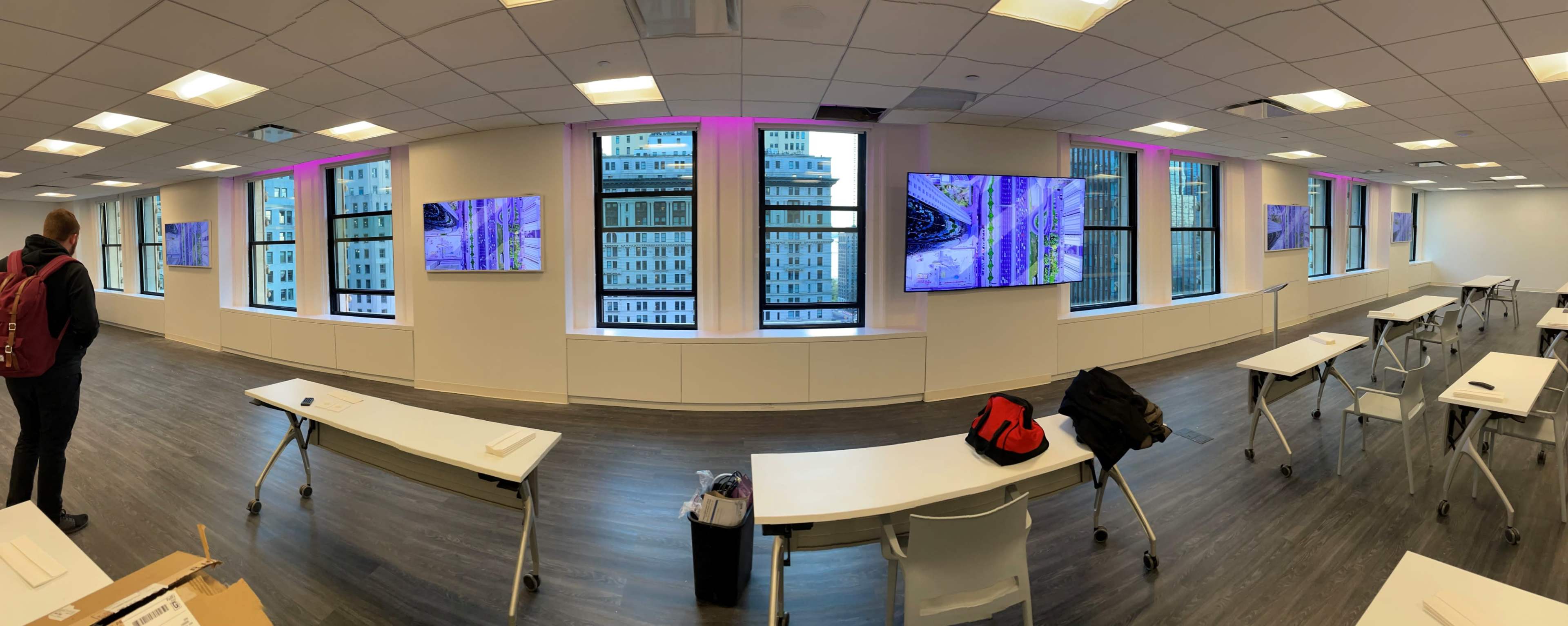 A spacious conference room with large windows, featuring several wall-mounted screens displaying images, and equipped with white tables and chairs.