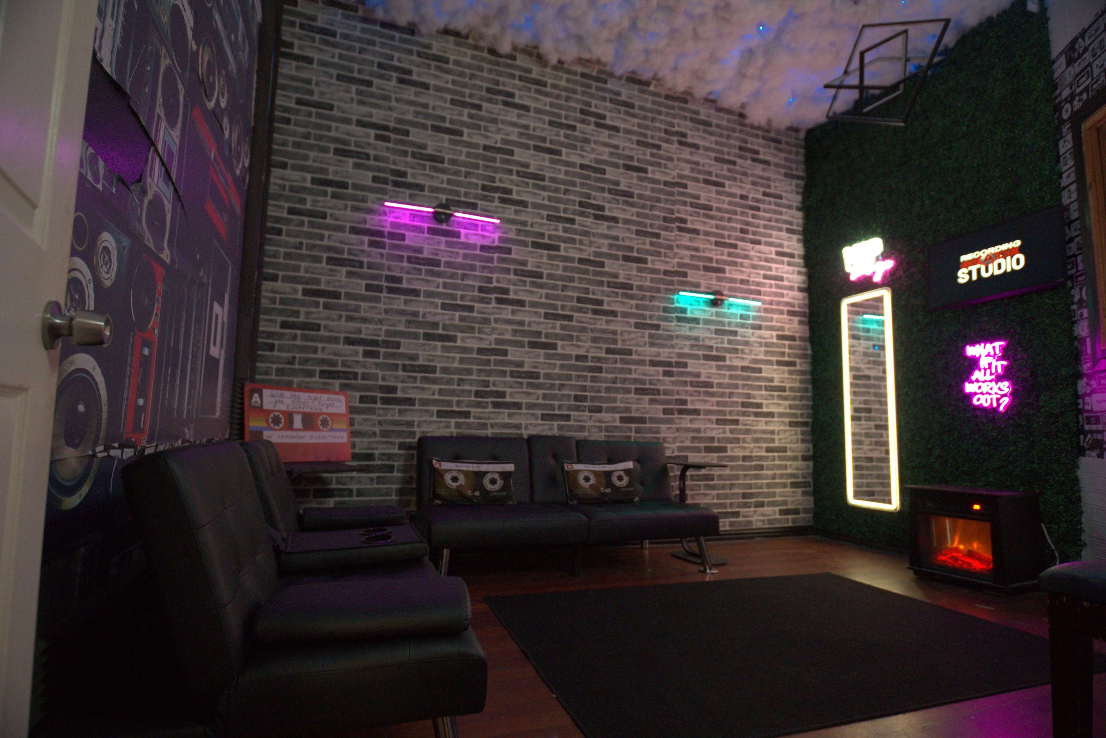 The image shows a modern lounge area with a brick wall, black leather sofas, neon lights, and a small electric fireplace.
