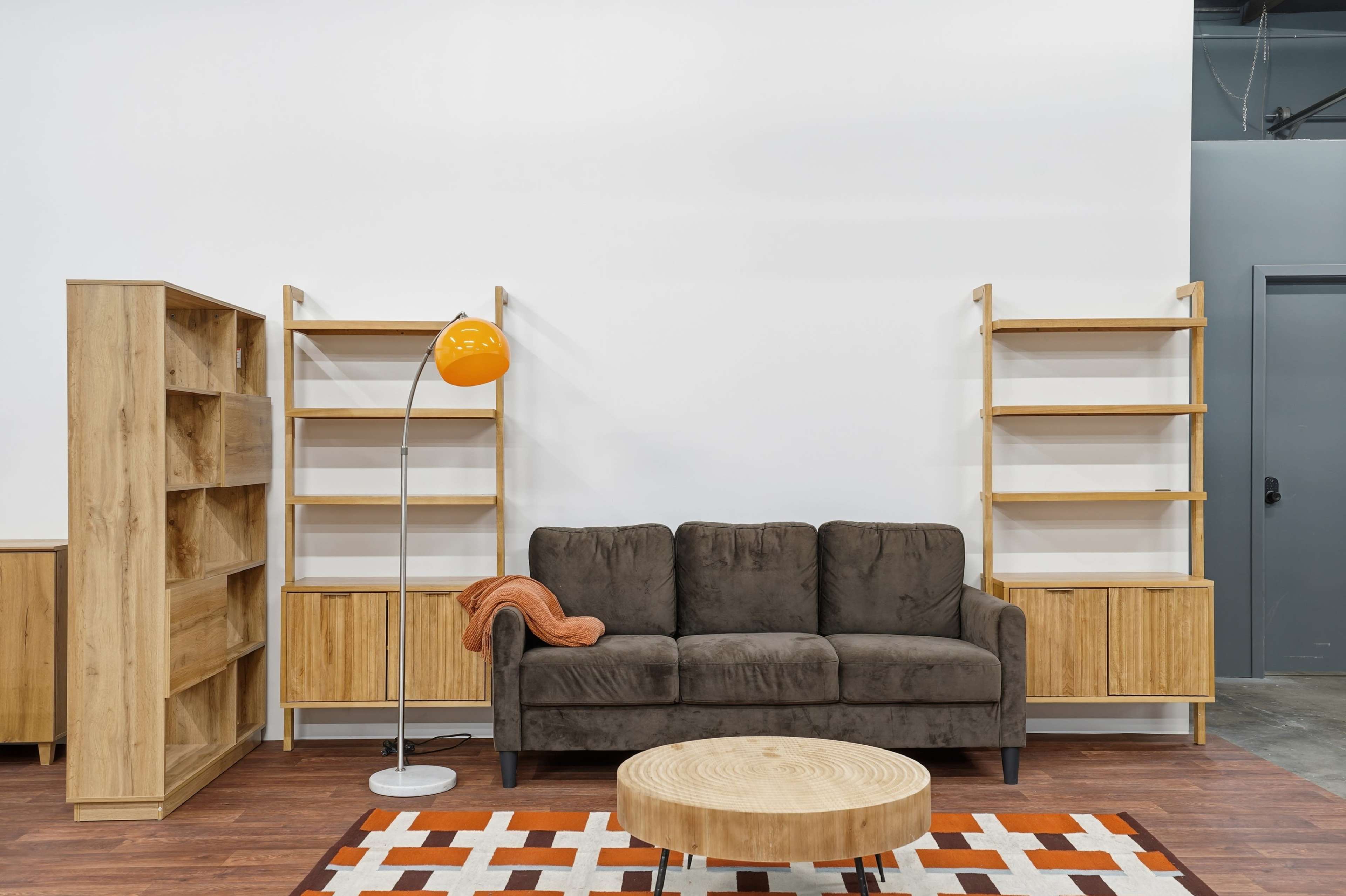 The image shows a modern living space featuring a brown sofa with an orange throw, a circular wooden coffee table, and wooden shelving units against a white wall.