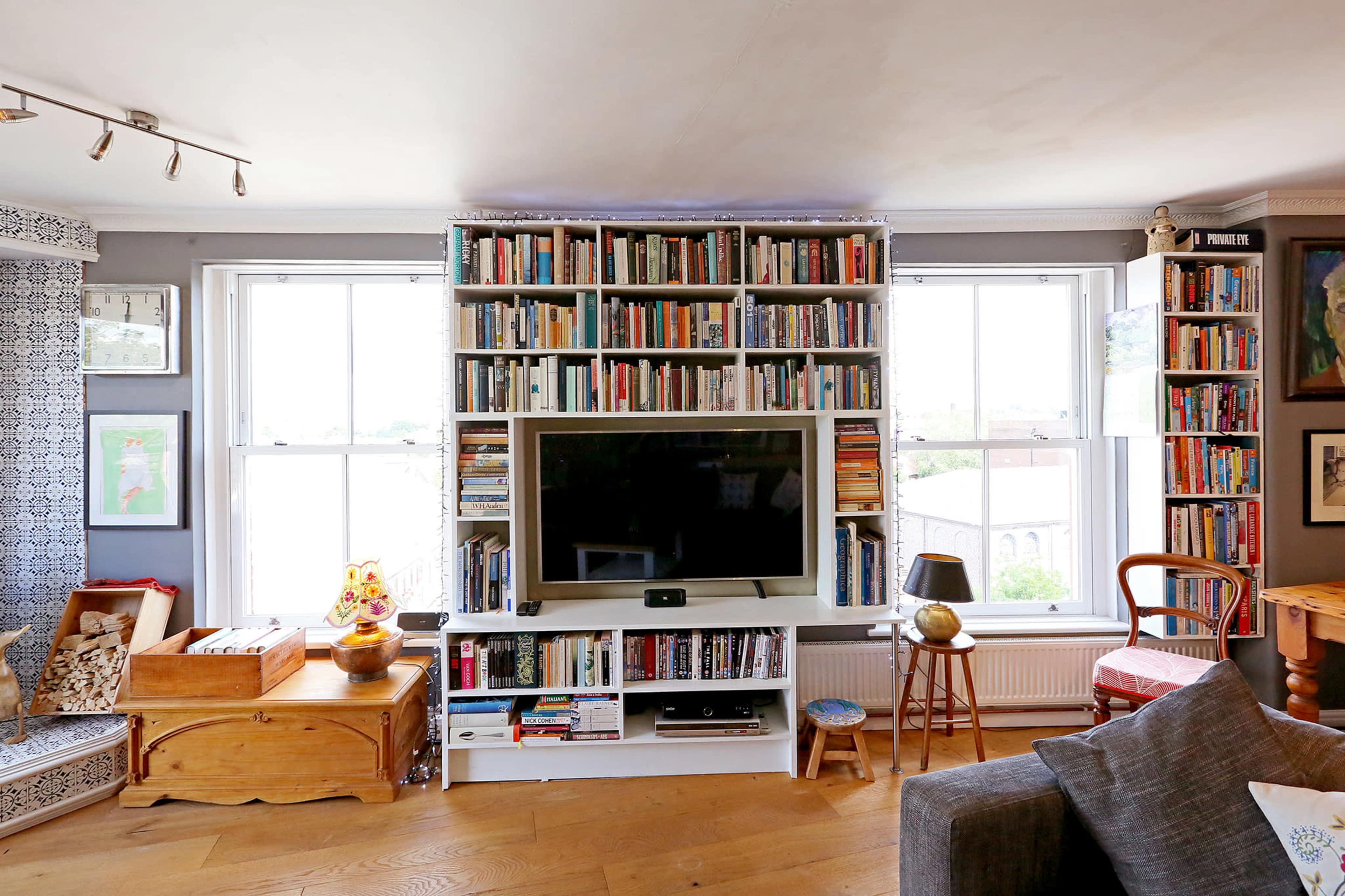 A cozy living room features a large bookshelf filled with books, a flat-screen TV, and windows that provide natural light.