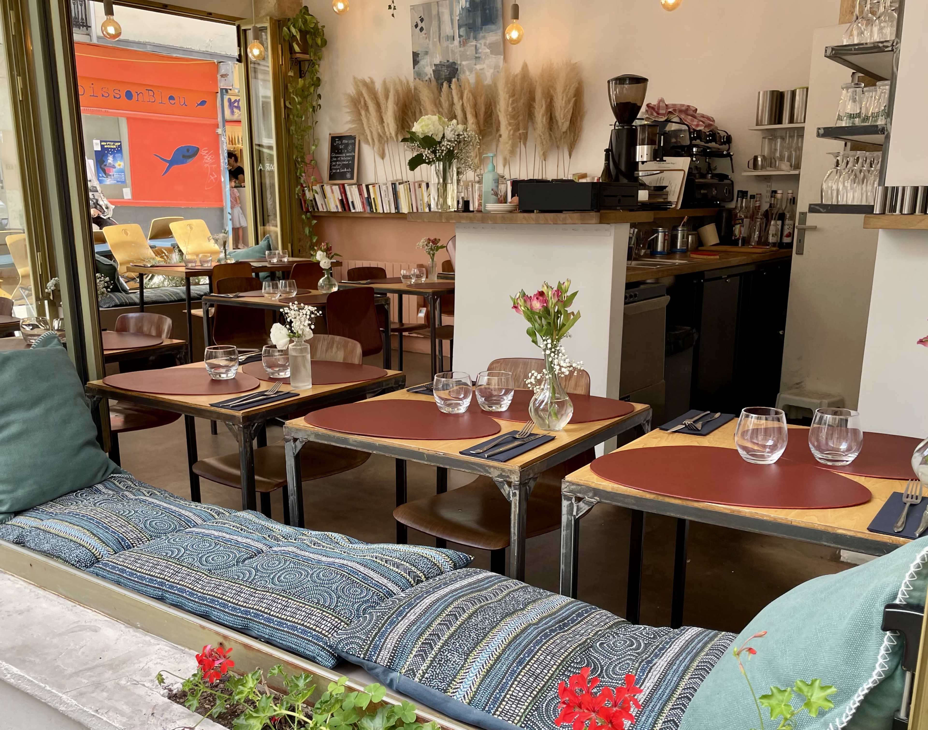 The image shows a cozy café interior with several tables set for dining, a coffee machine on the counter, and decorative plants in the background.
