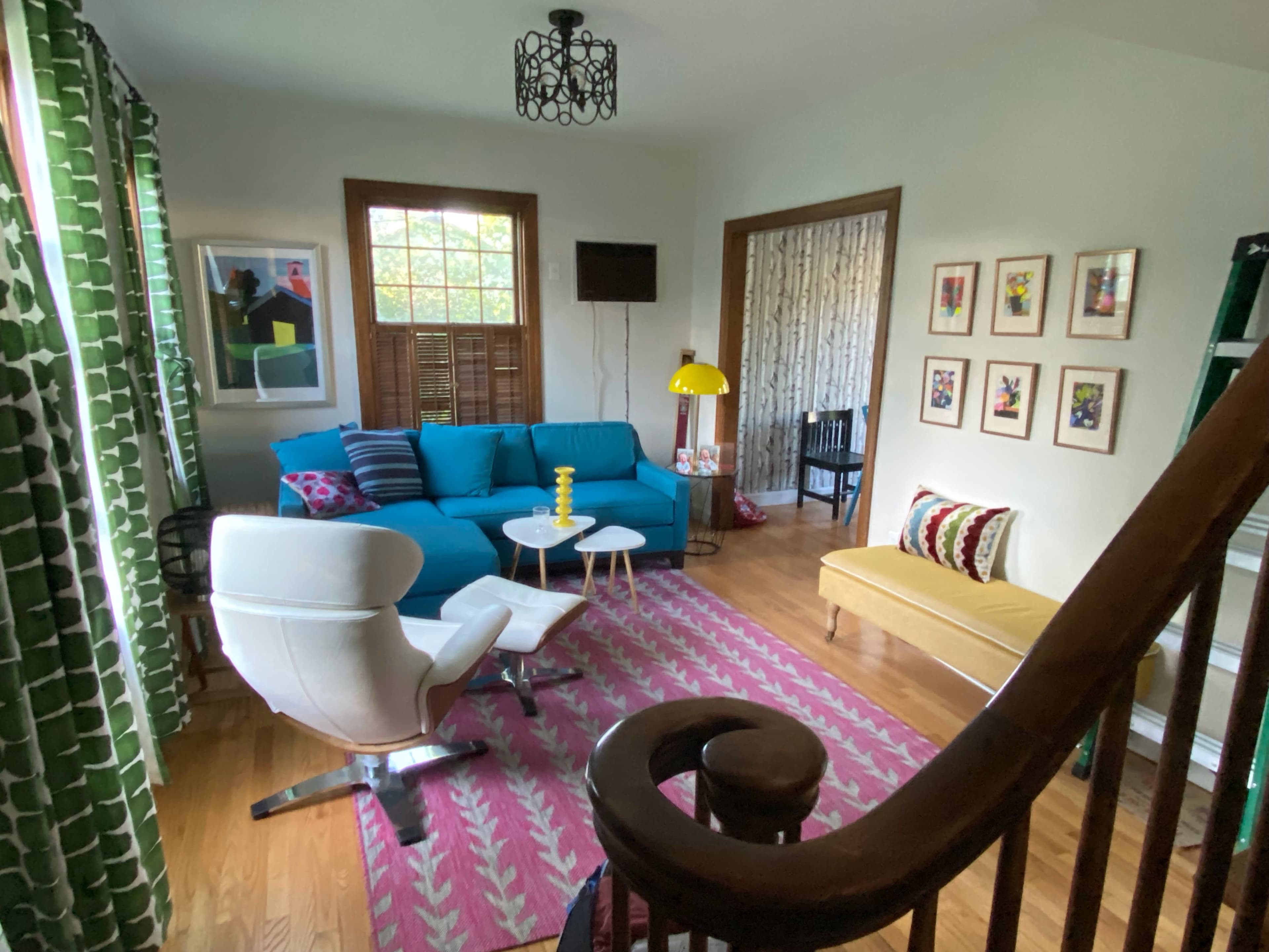 A bright living room features a blue sofa, a yellow bench, patterned curtains, and various framed artworks on the walls.
