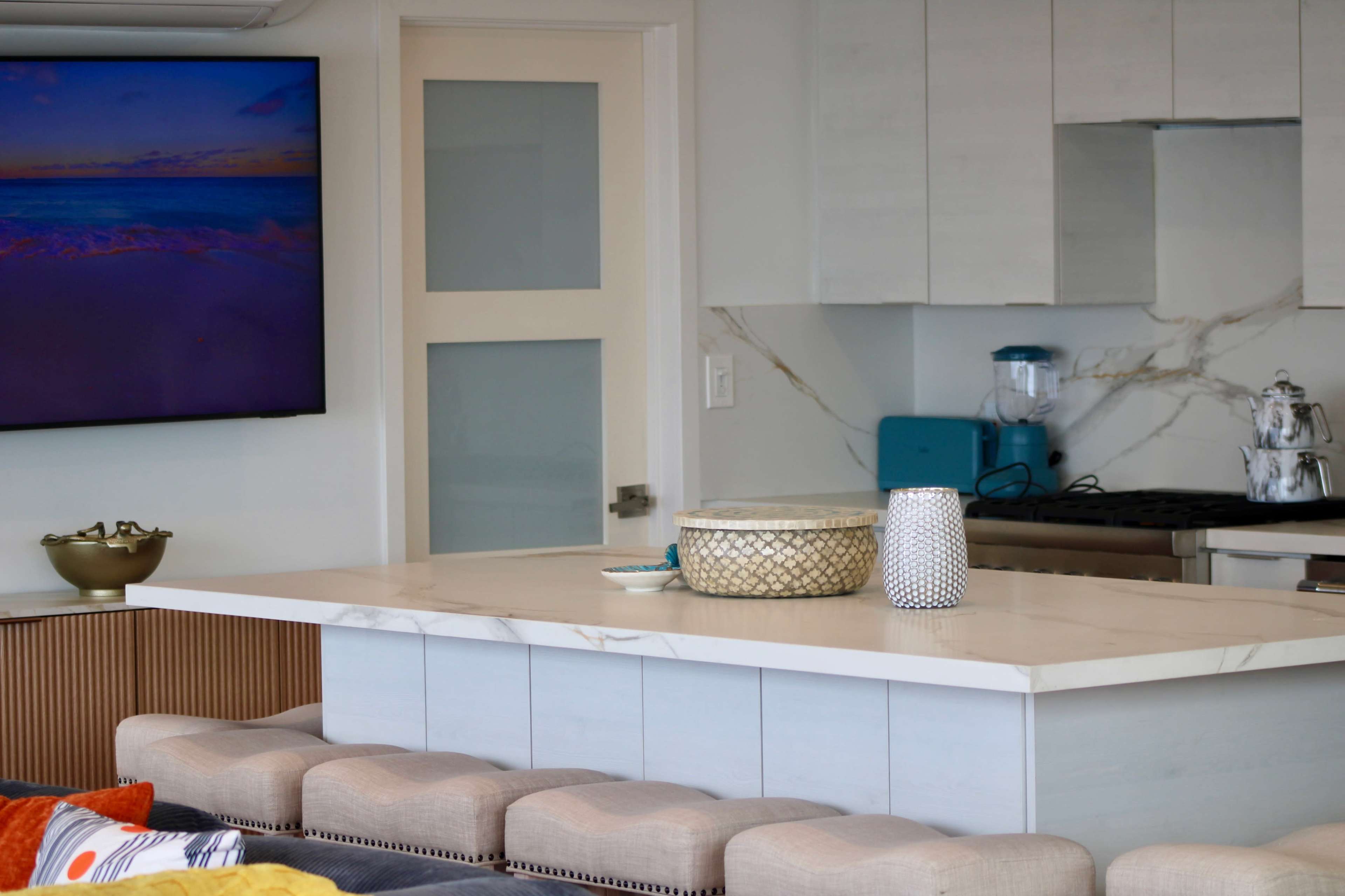 The image shows a modern kitchen and living area featuring a quartz countertop, a television mounted on the wall, and a blue kitchen appliance.