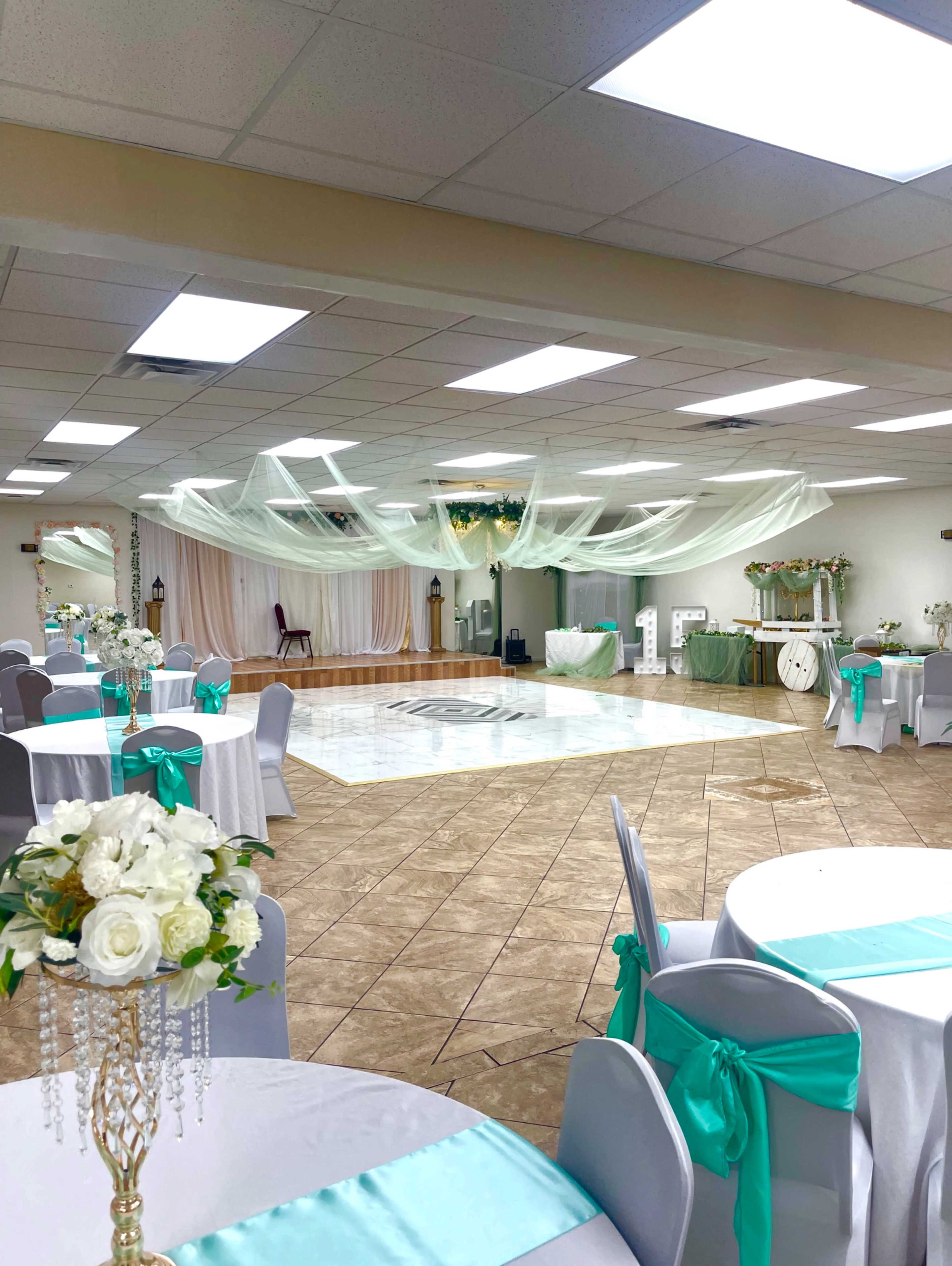 A spacious event hall features decorated tables with turquoise accents, elegant draping overhead, and a central dance floor.