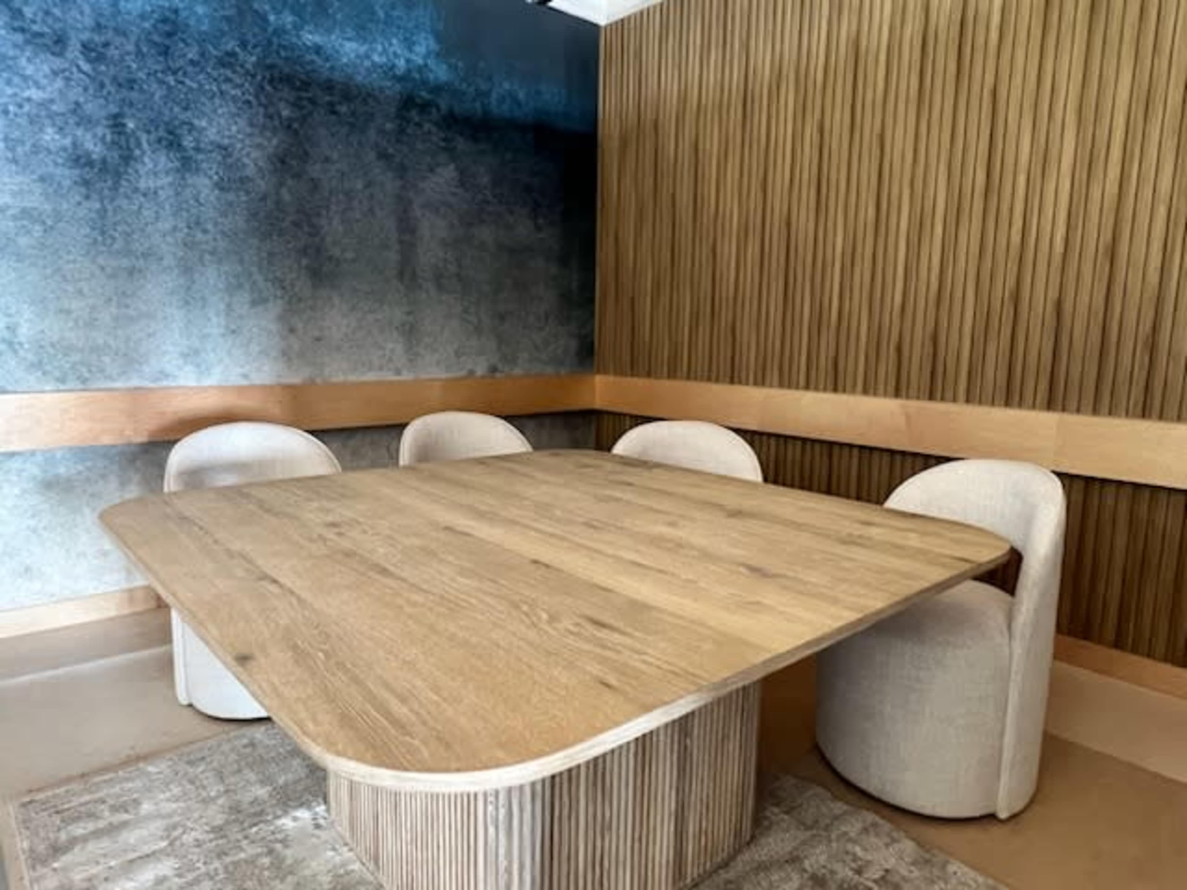 The image shows a modern conference room with a wooden table and four upholstered chairs against a backdrop of textured blue and wooden walls.