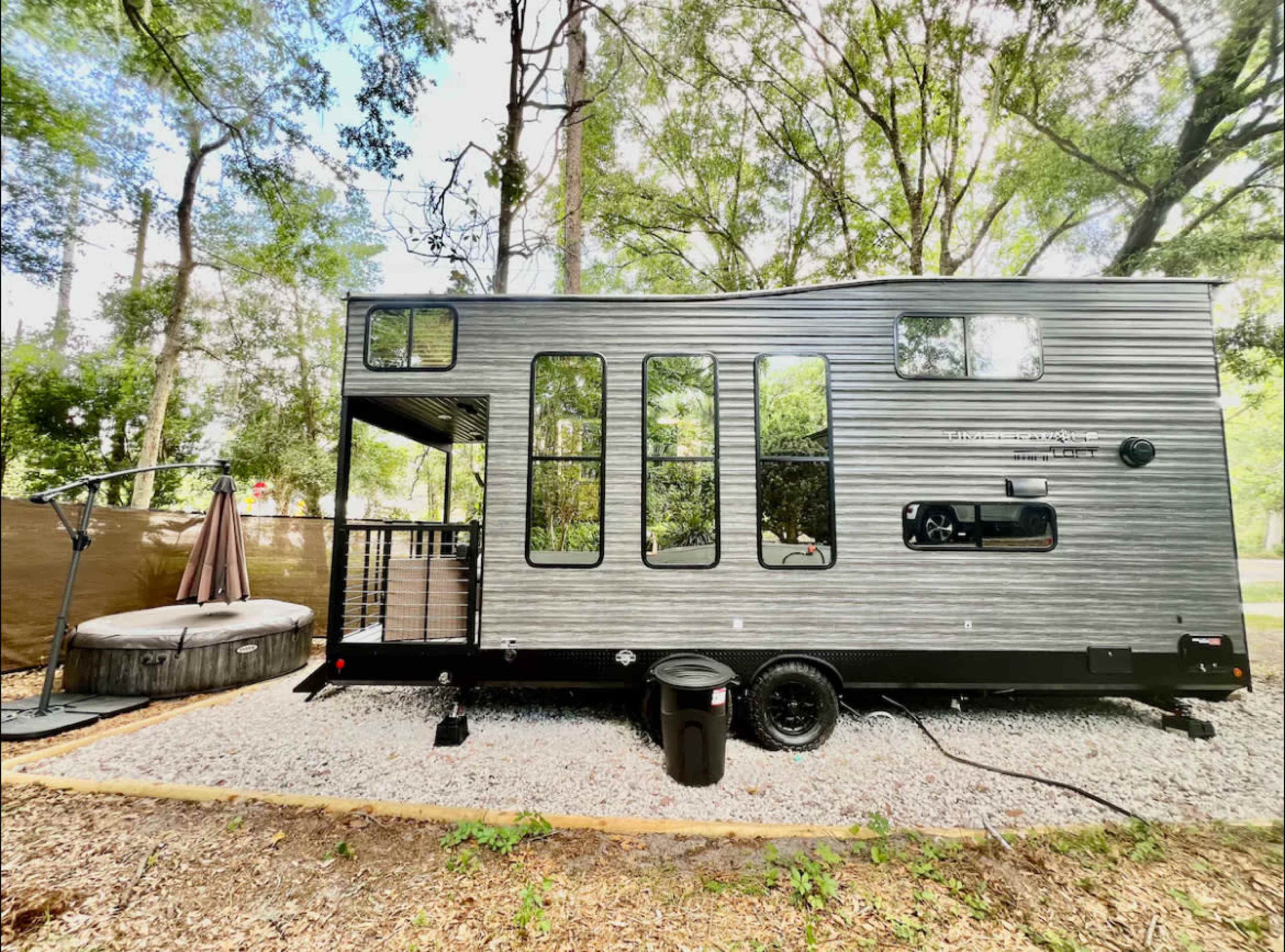 A modern travel trailer with multiple windows is parked on gravel beside a hot tub in a wooded area.