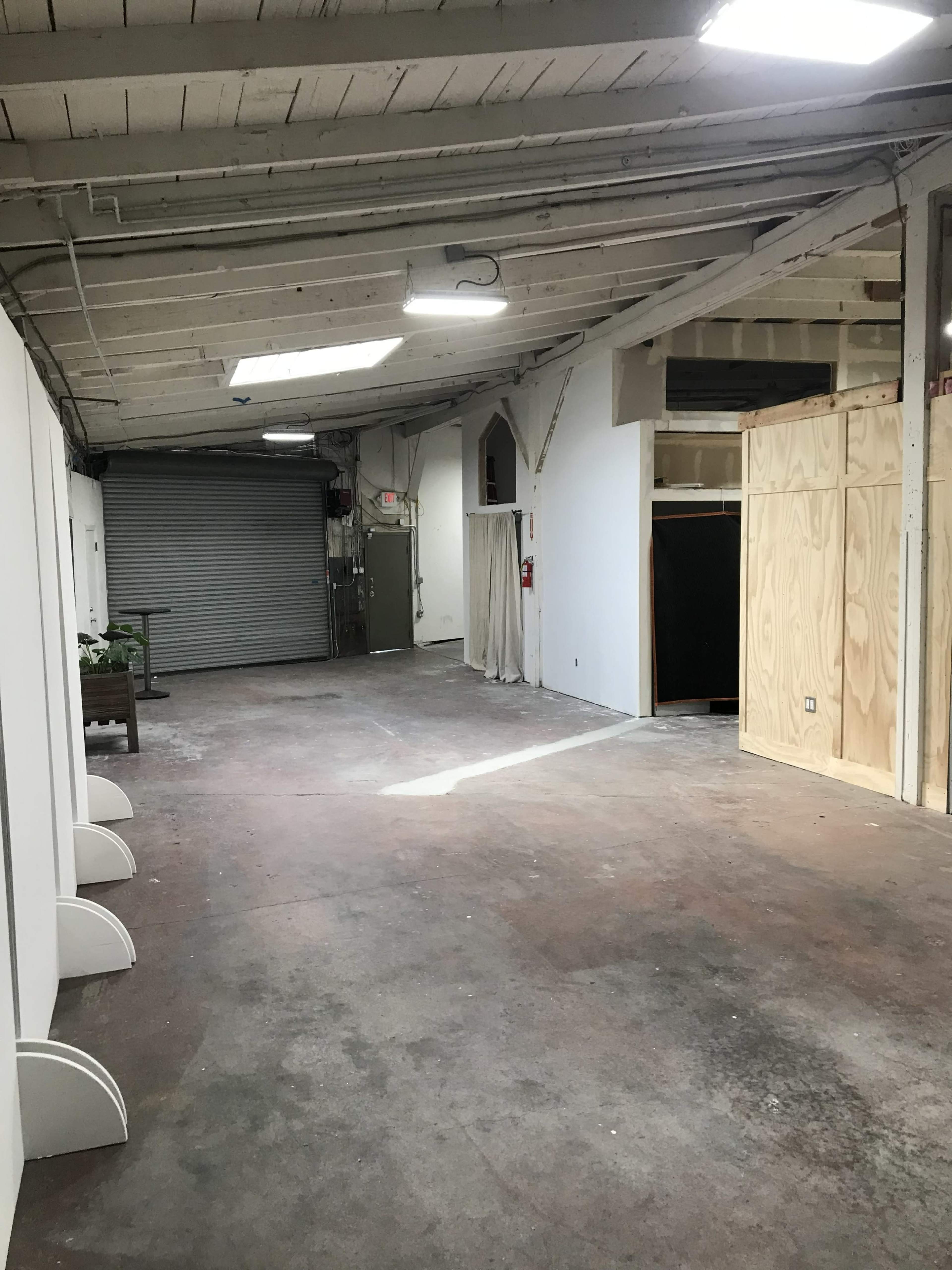 The image shows an empty industrial space with exposed beams, a metal door on one side, and light streaming in from a skylight.