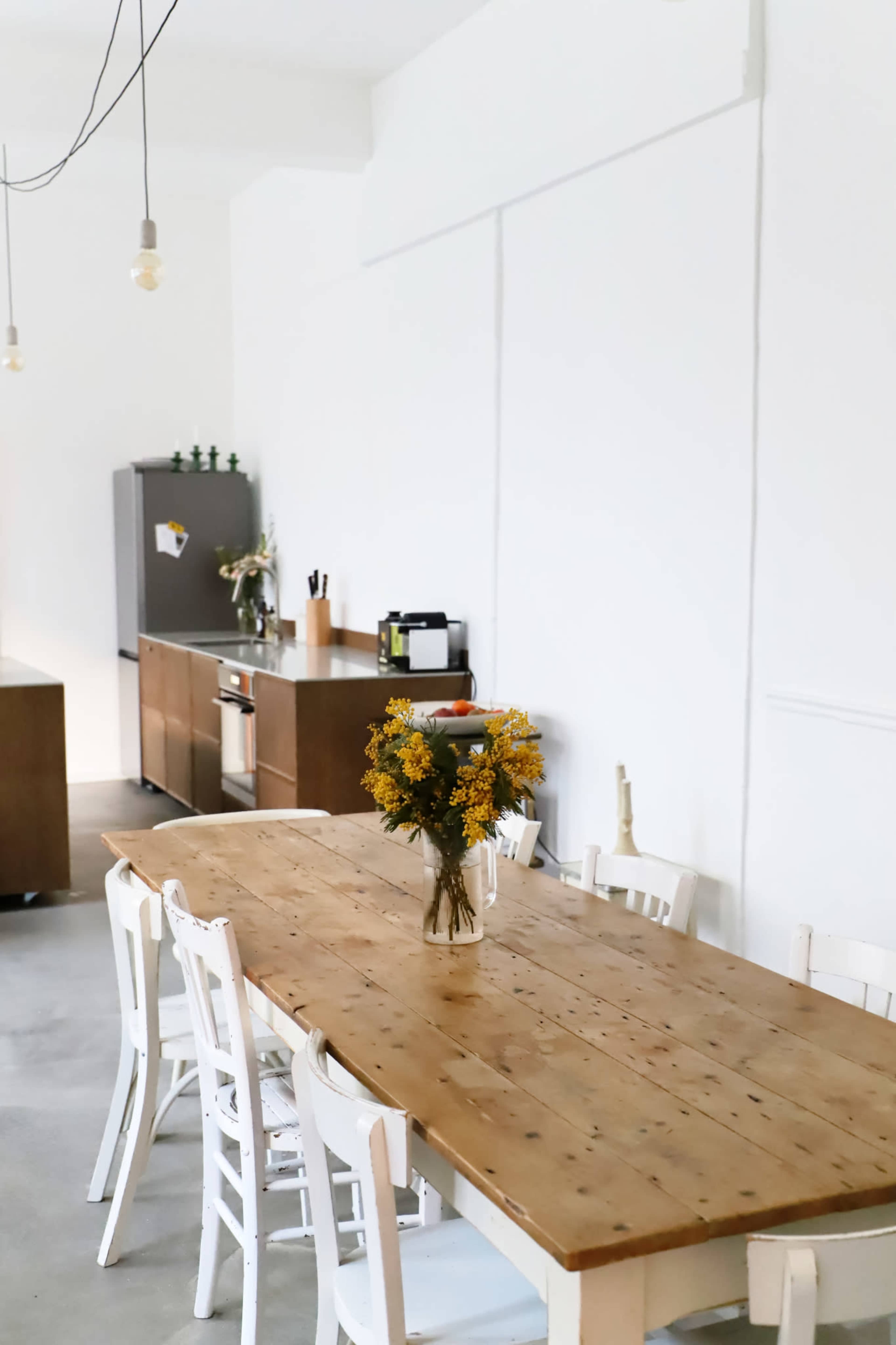 A wooden table with a vase of yellow flowers sits in a minimalist kitchen featuring white walls and modern appliances.