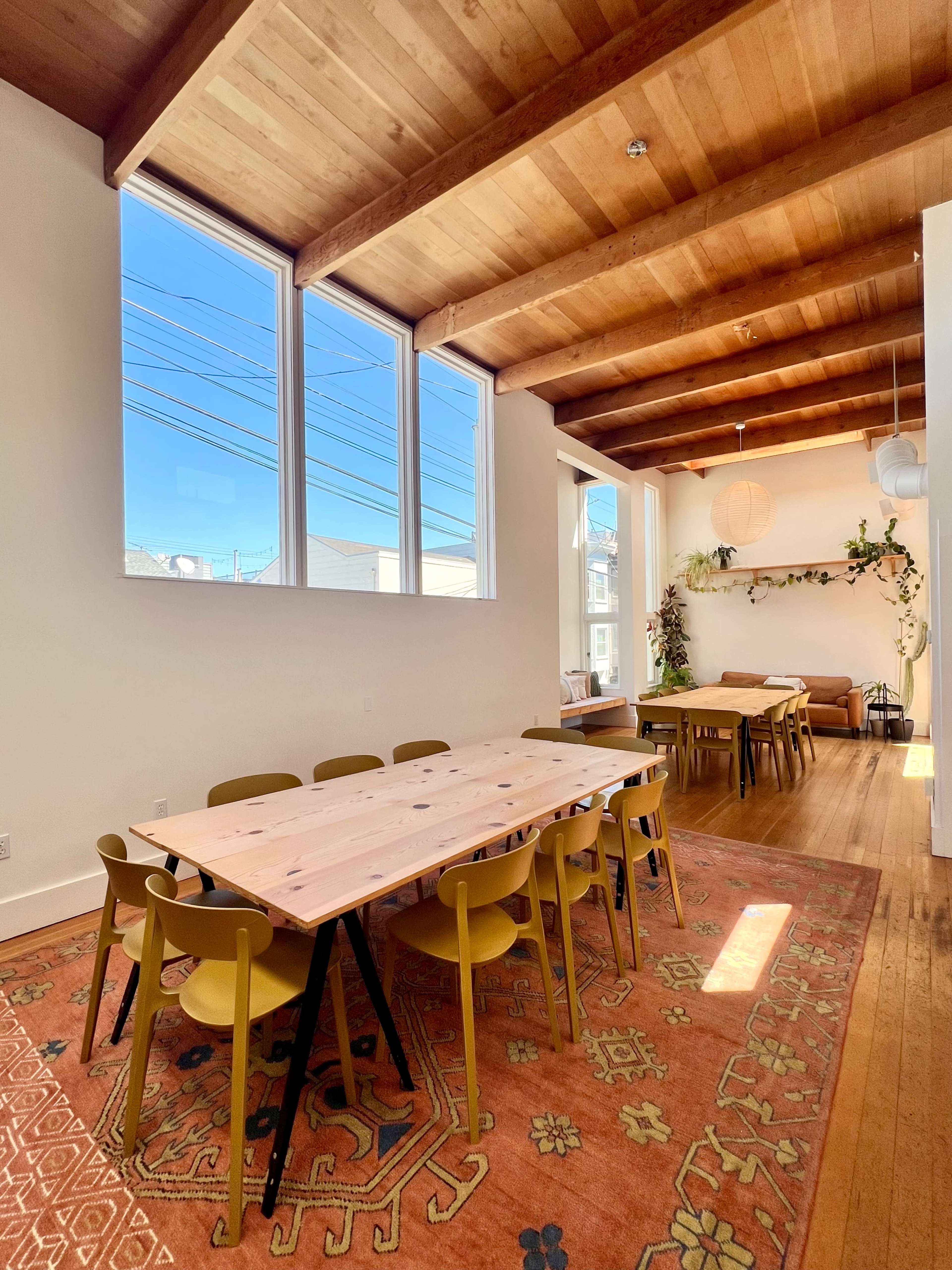 The image shows a bright, airy room with wooden beams, large windows, a large area rug, and multiple tables surrounded by chairs.