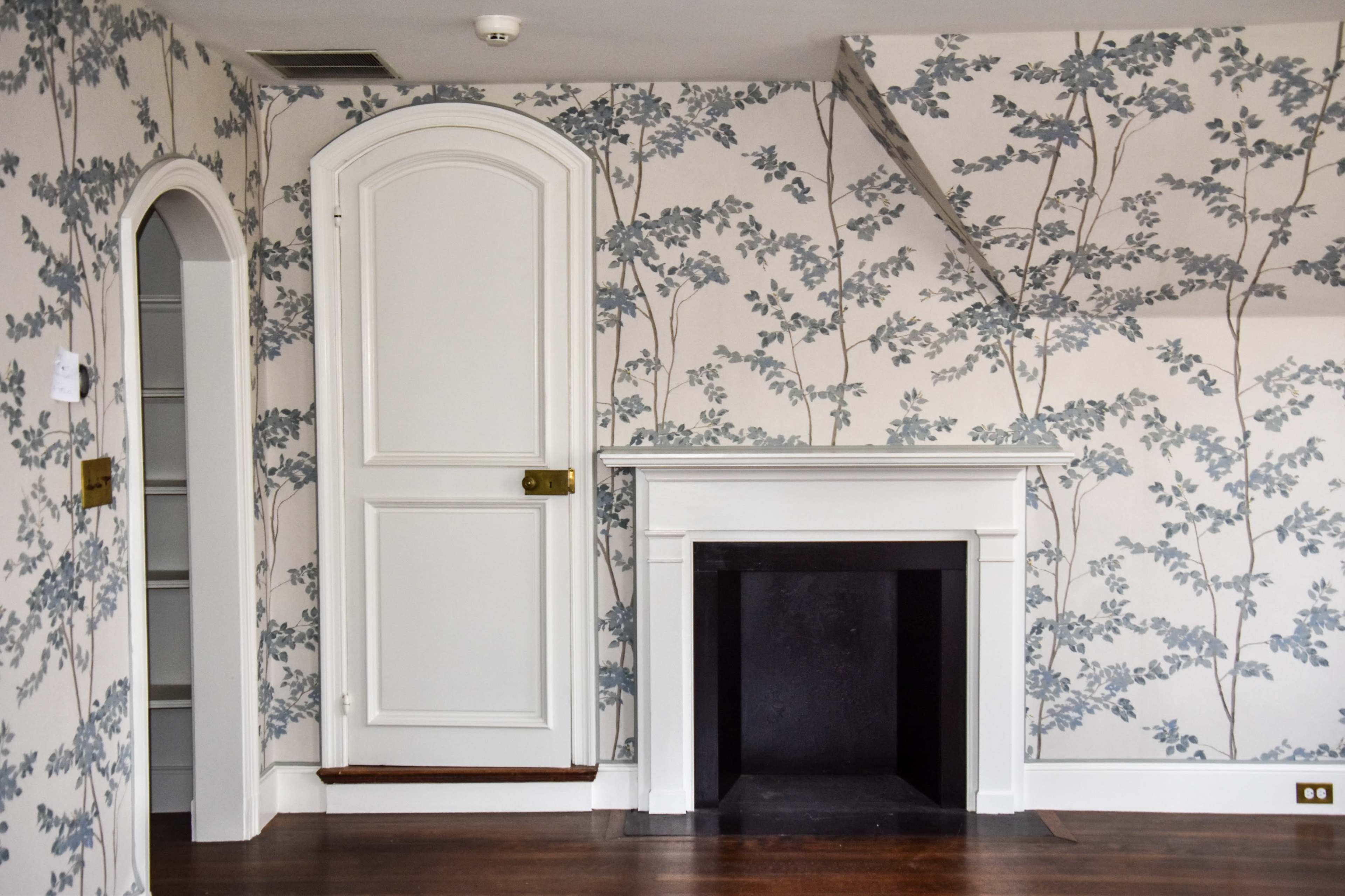 A room with floral wallpaper, a closed door, a fireplace without a fire, and hardwood flooring.