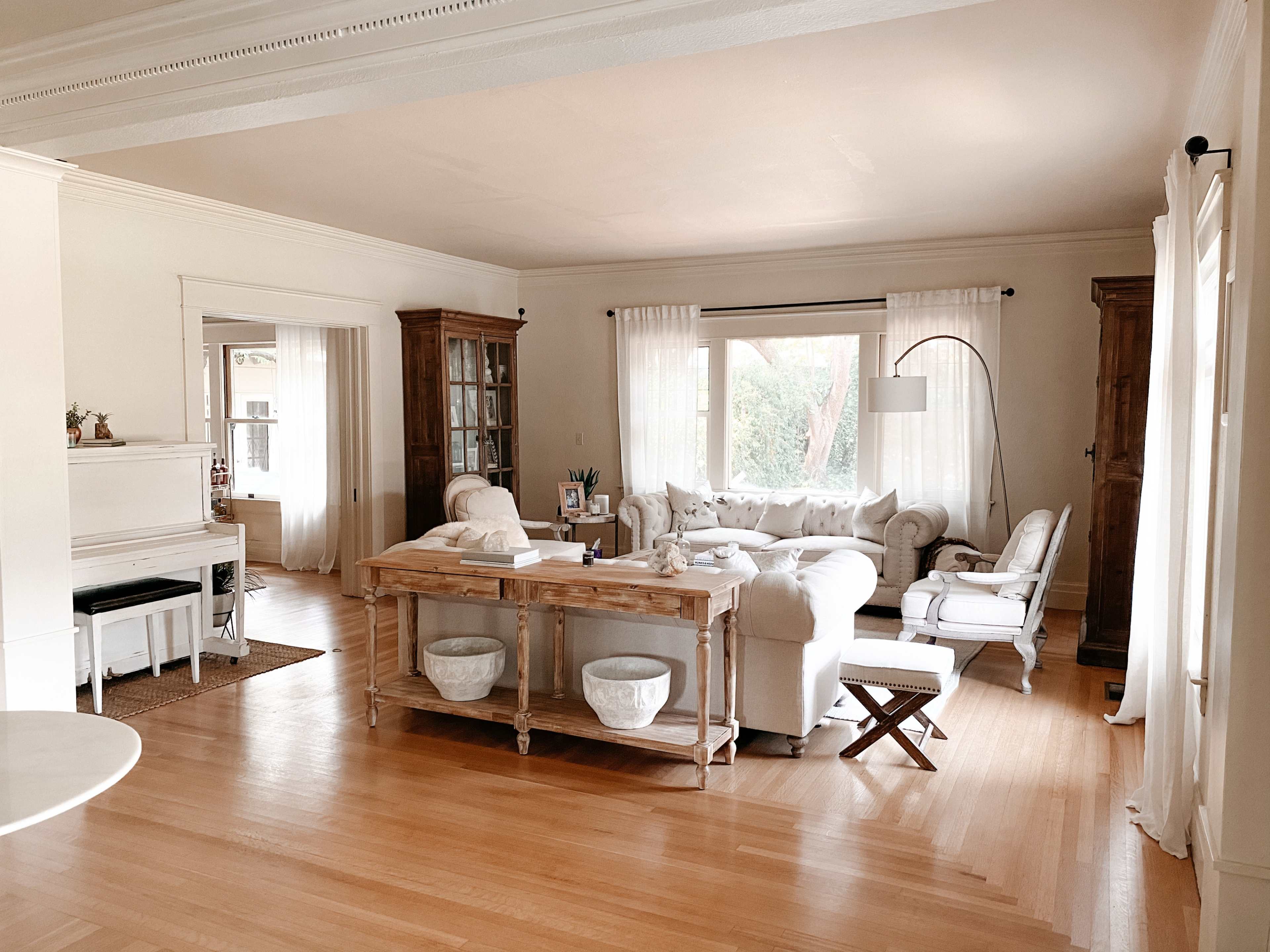 A bright living room features a light-colored sofa, a wooden coffee table, and large windows with sheer curtains.