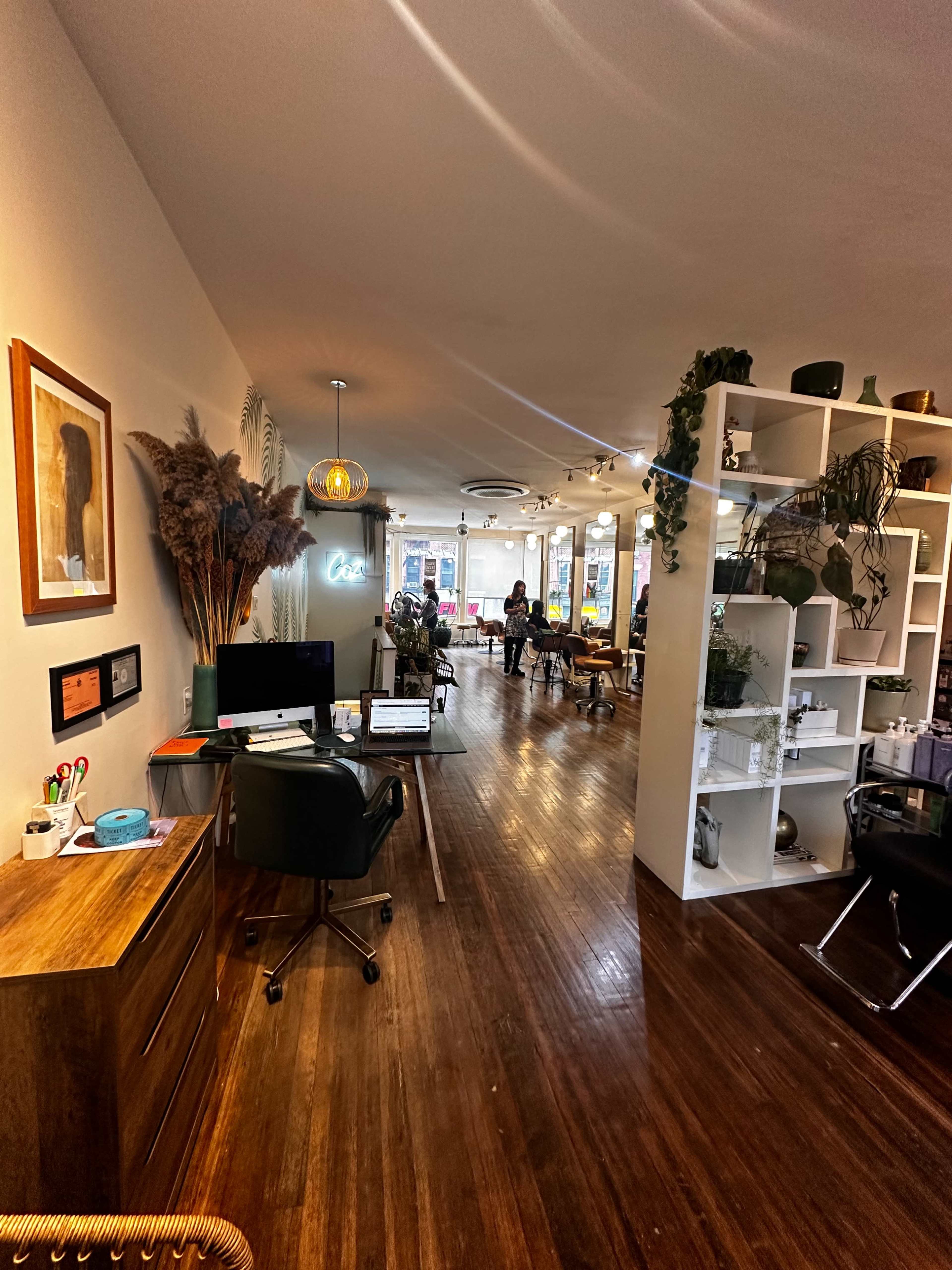 The image shows an interior of a modern, open-concept workspace with wooden flooring, plants, and a reception area.