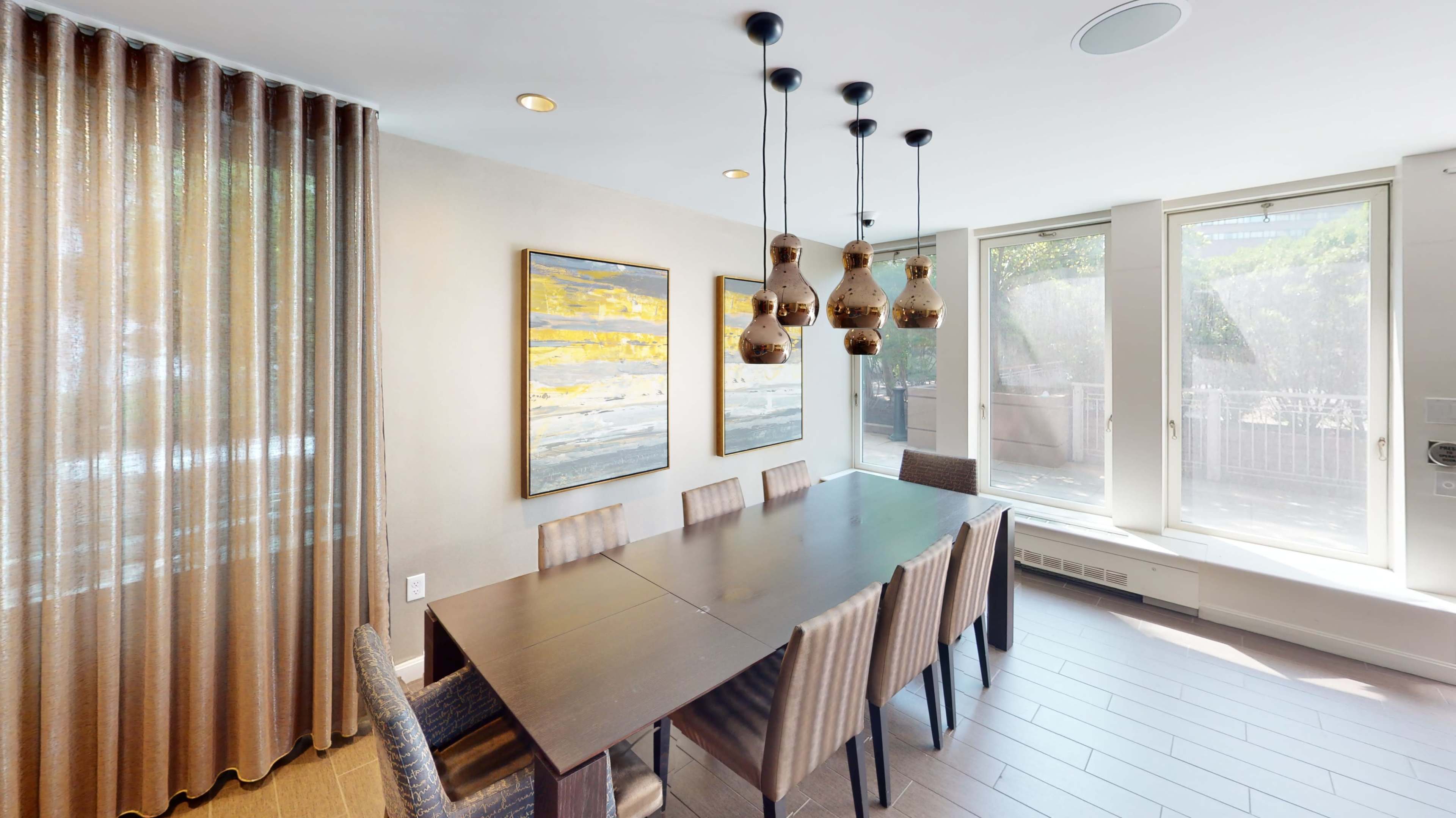 A dining area features a long table surrounded by chairs, with pendant lights overhead and large windows letting in natural light.