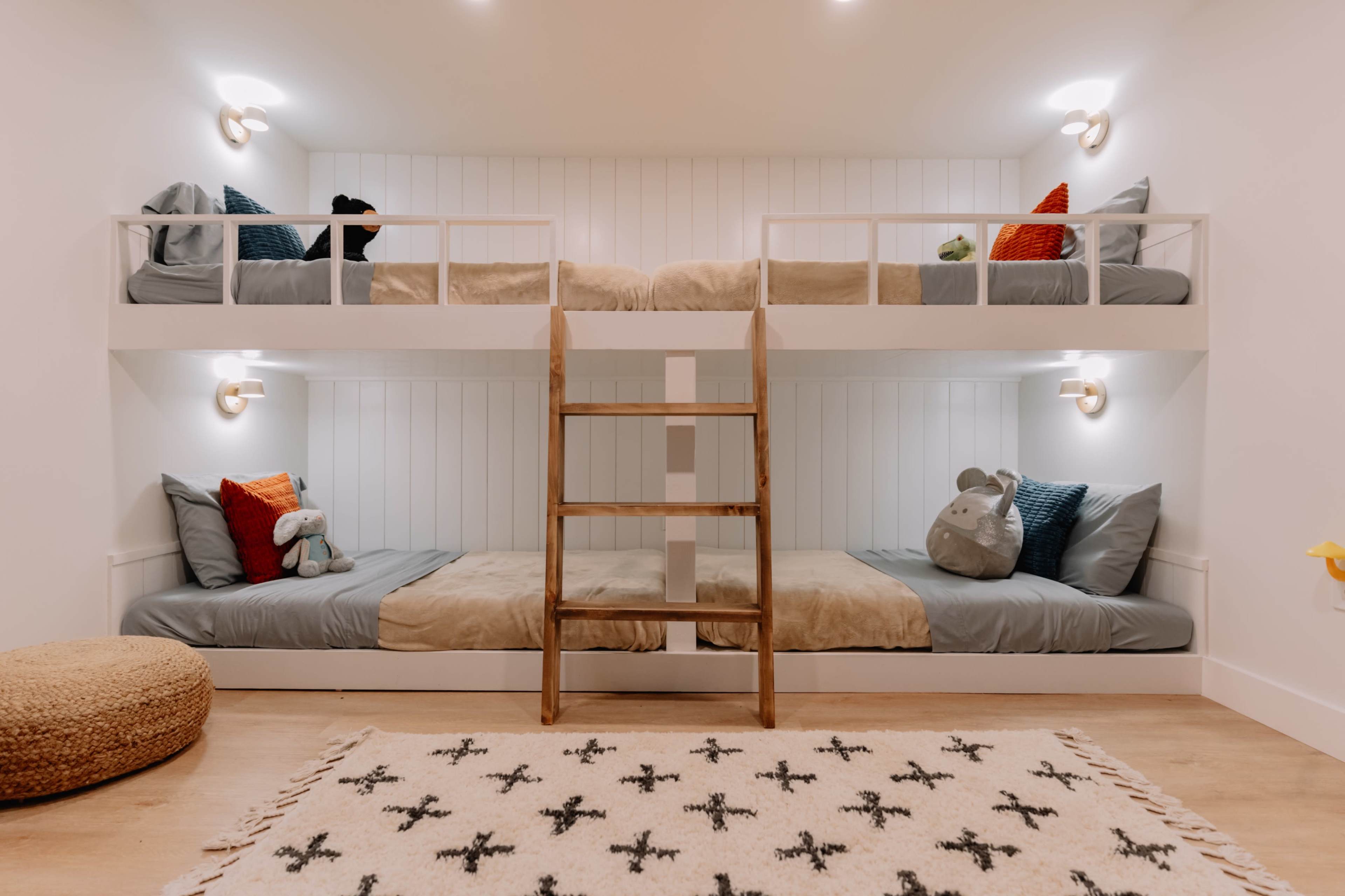 The image shows a bedroom with two sets of wooden bunk beds arranged in a cozy layout, featuring simple bedding and soft toys on each bed.