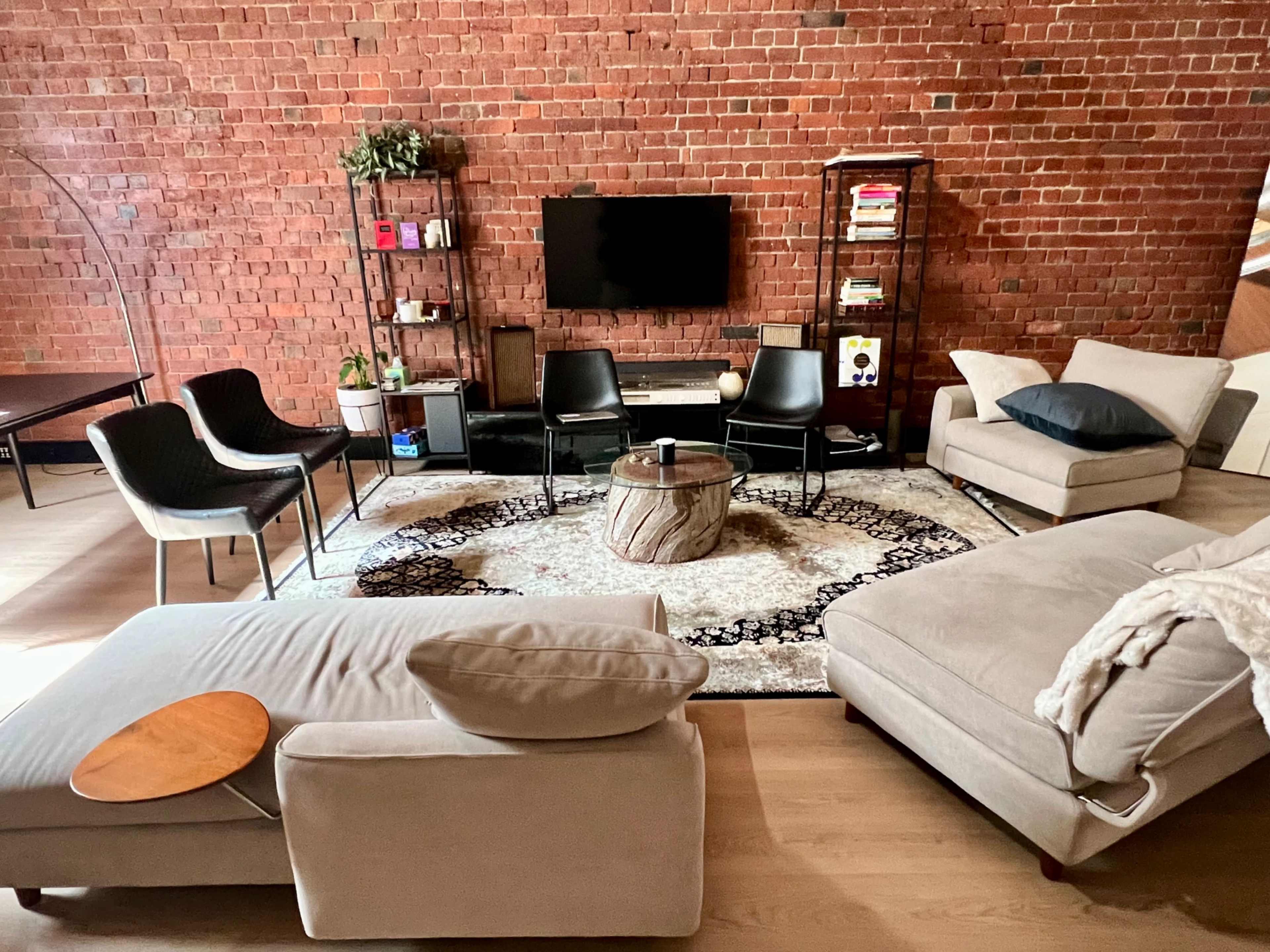 The image shows a modern living room with exposed brick walls, a circular rug, a television mounted on the wall, and various seating options arranged for relaxation.