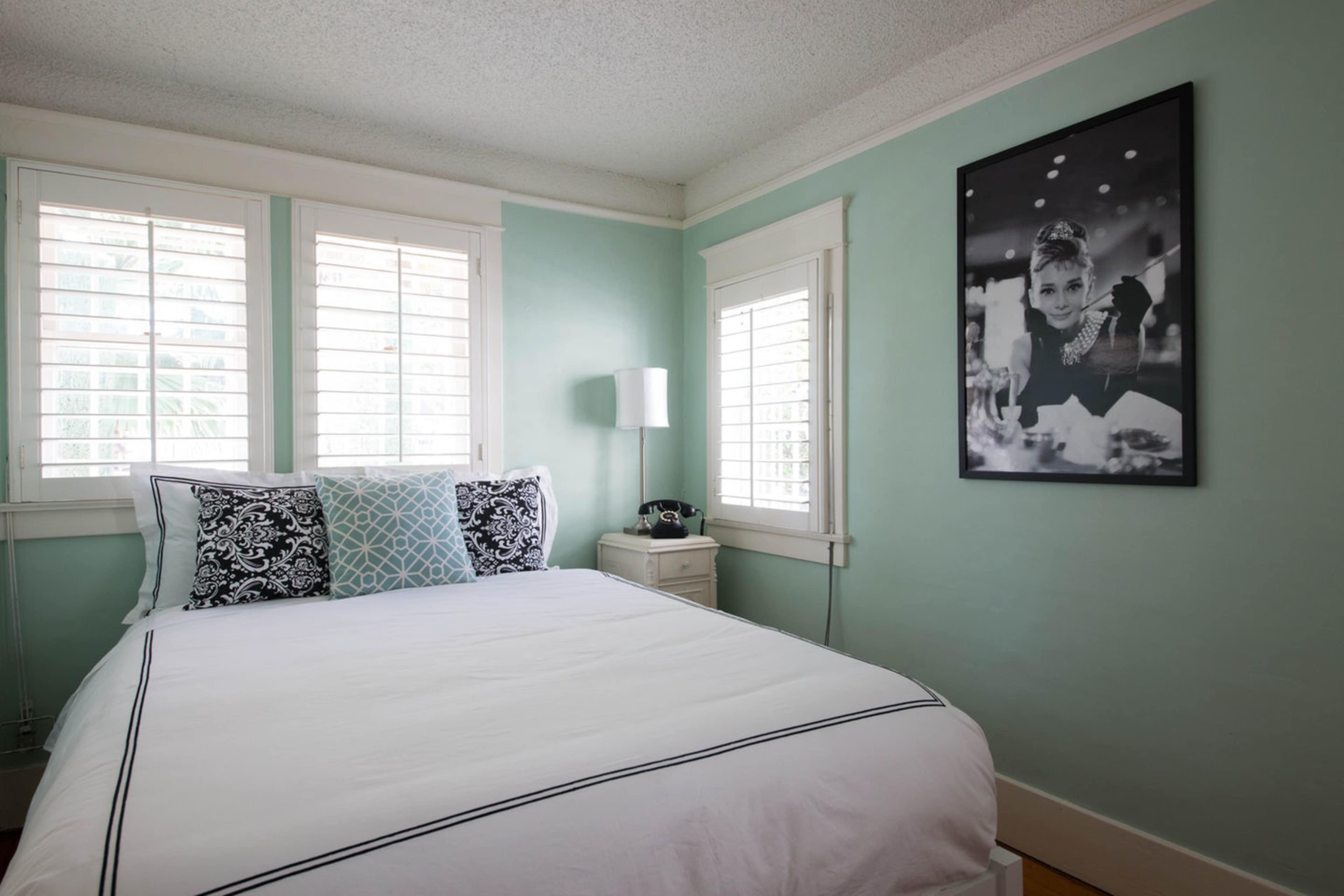 A light green bedroom features a bed with a white duvet and black accents, accompanied by a large black-and-white photograph on the wall.